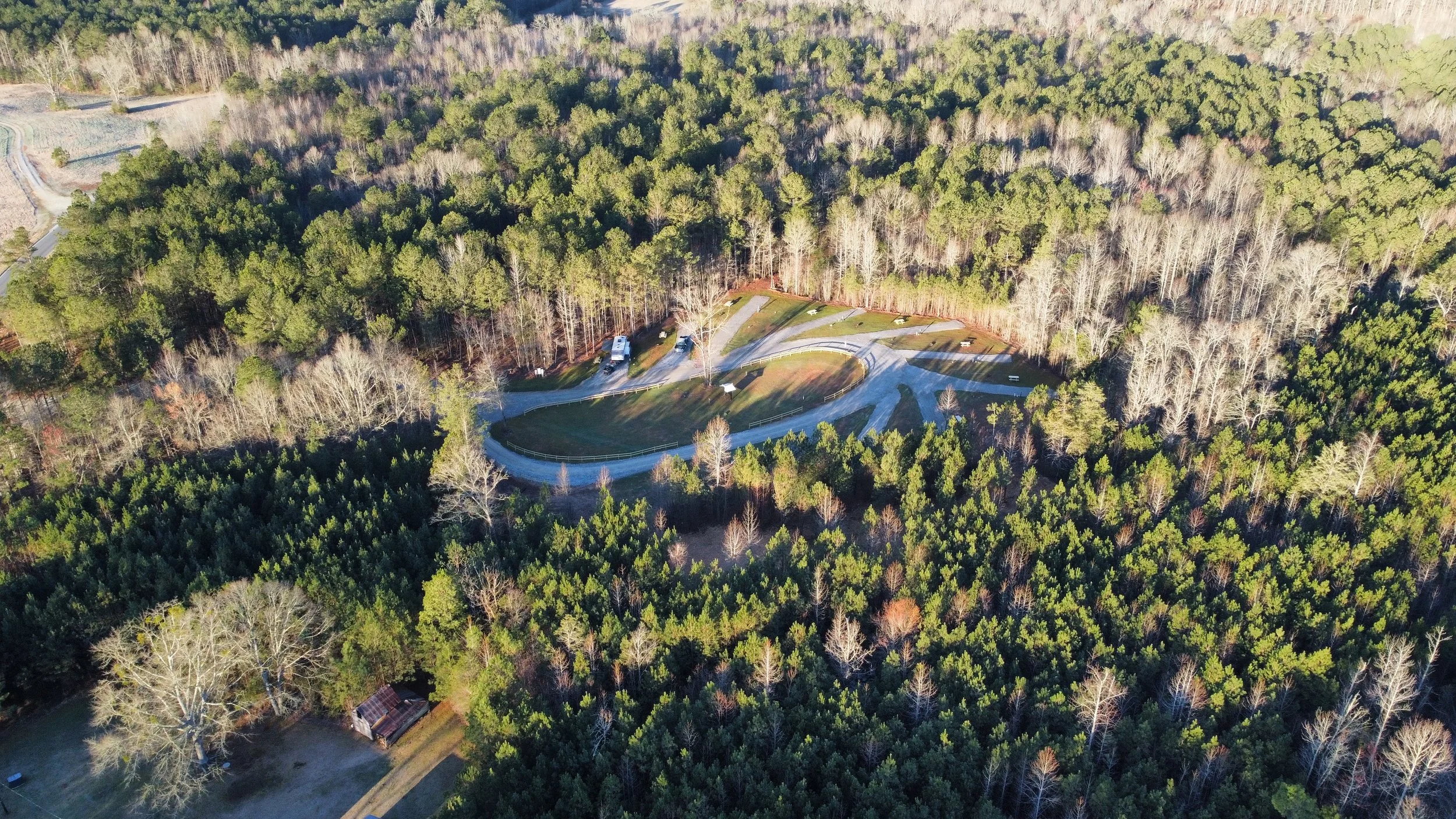 An aerial view of a forested area with a small parking lot, several vehicles, and a gravel road. The trees are mostly deciduous with some evergreen trees.