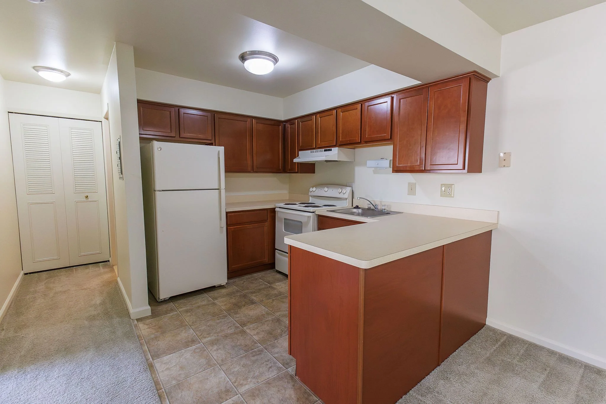 Kitchen with white refrigerator, oven, and cabinets with wood finish, beige countertops, and a small breakfast bar, adjacent to a beige carpeted area.