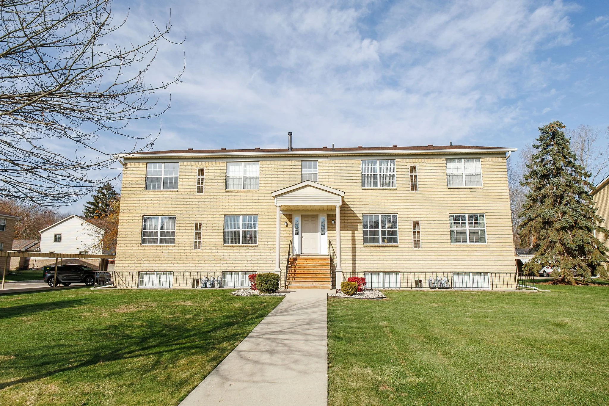 A beige multi-unit residential building with a central staircase and front door, surrounded by a green lawn and trees, under a partly cloudy sky.