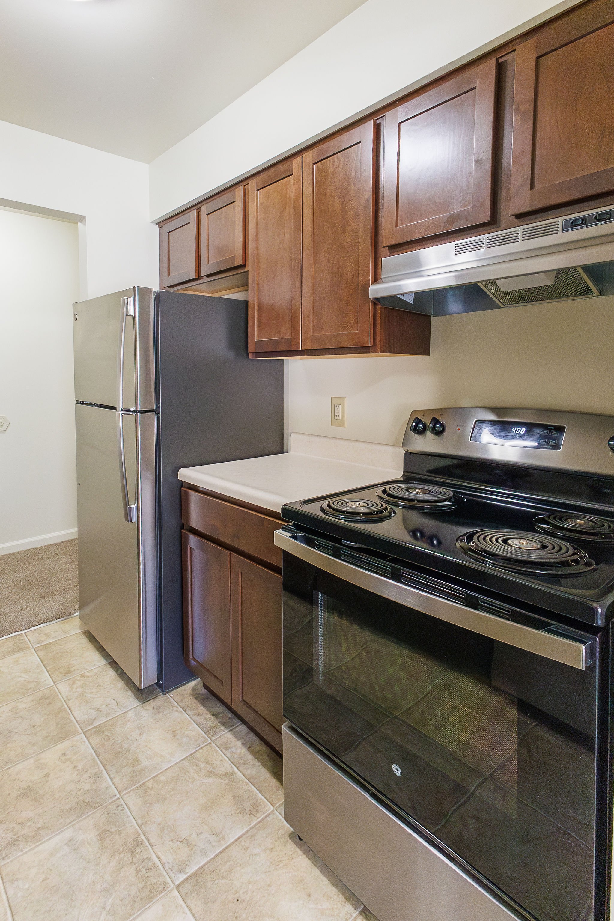 A kitchen with a stainless steel refrigerator, wooden cabinets, a beige countertop, and a black electric stove.