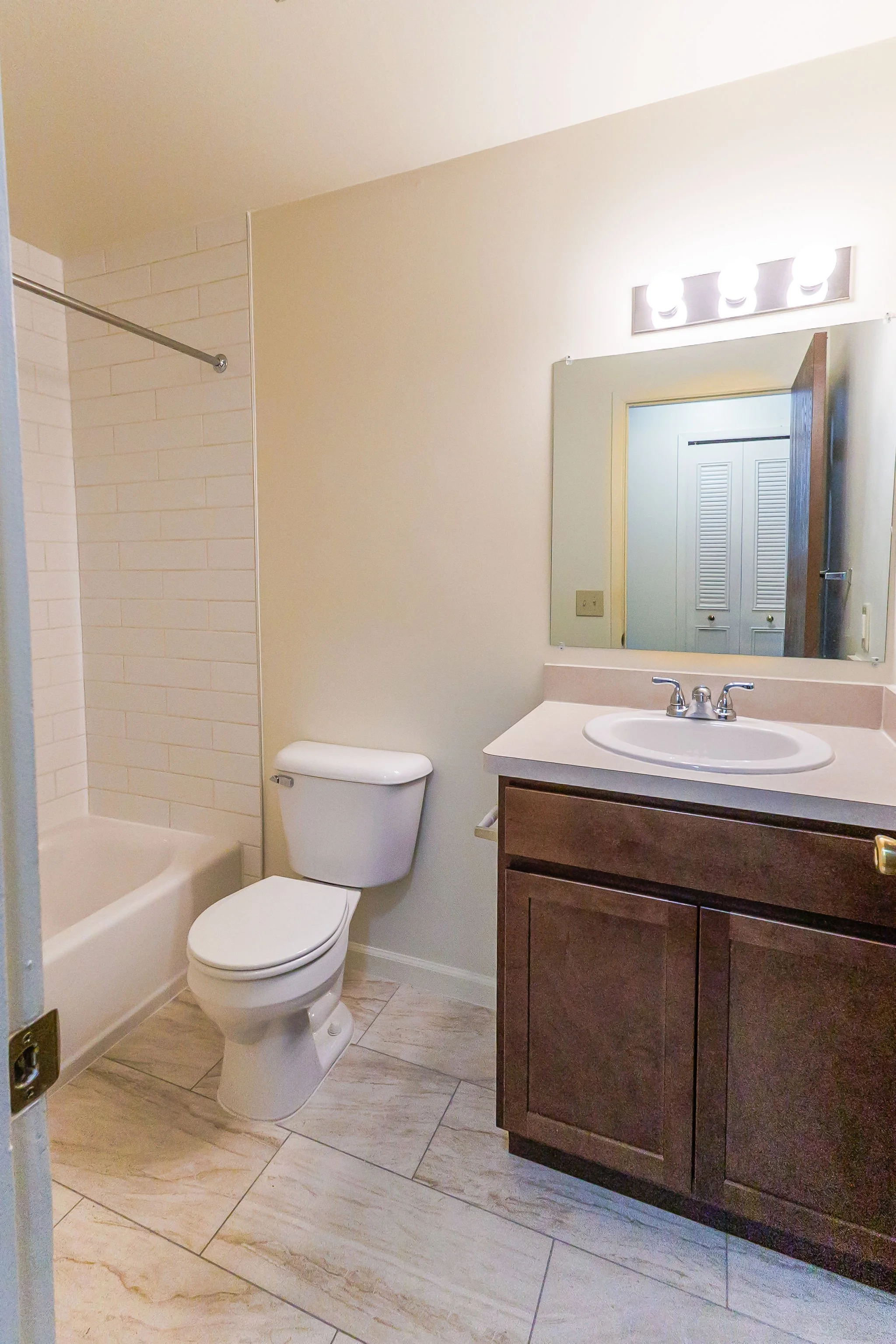 A small bathroom with a bathtub and shower, a toilet, a wooden vanity with a sink, a mirror, and a row of lights above the mirror.