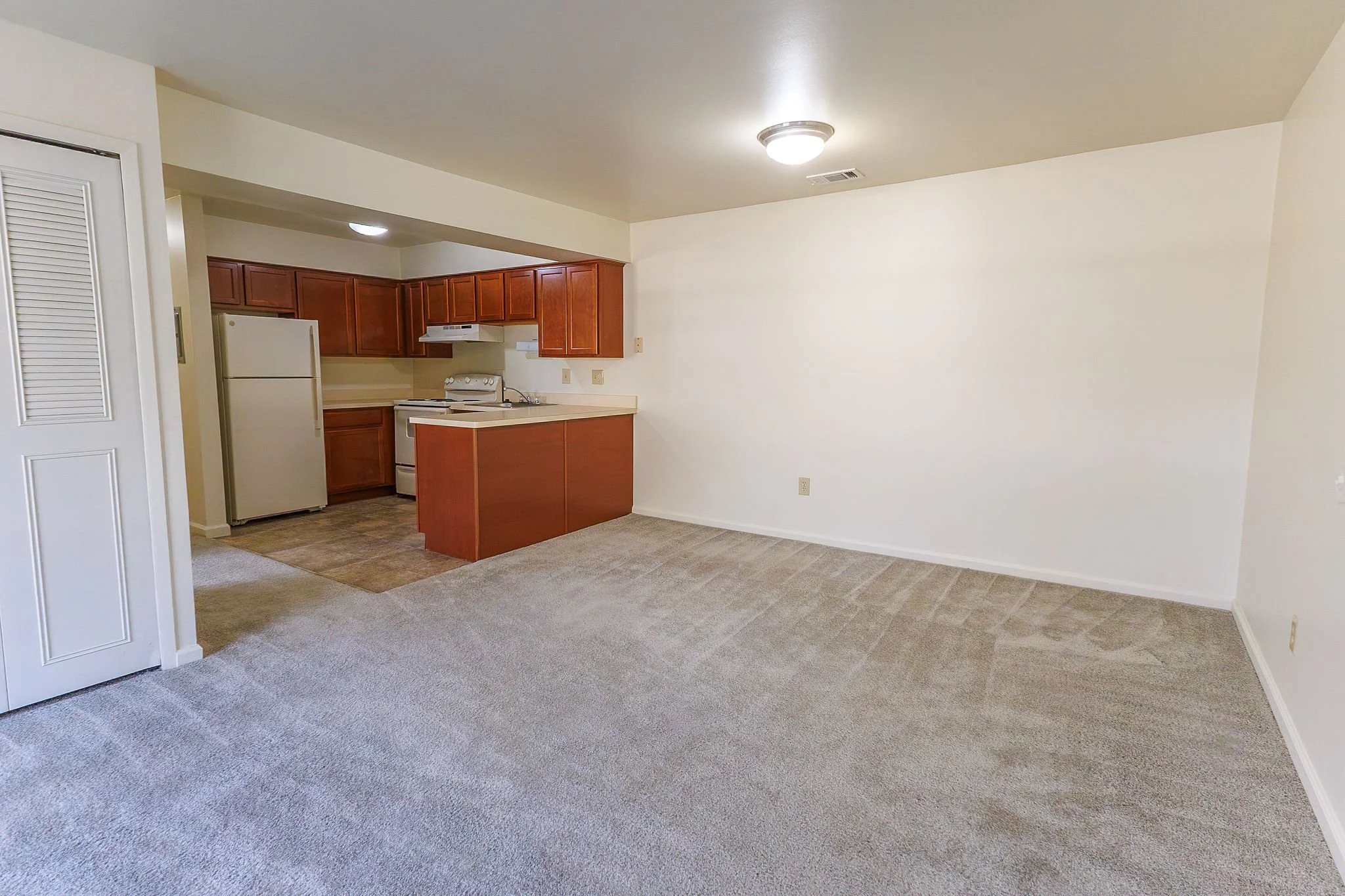 Empty living room with beige carpet and open kitchen with wooden cabinets and white appliances