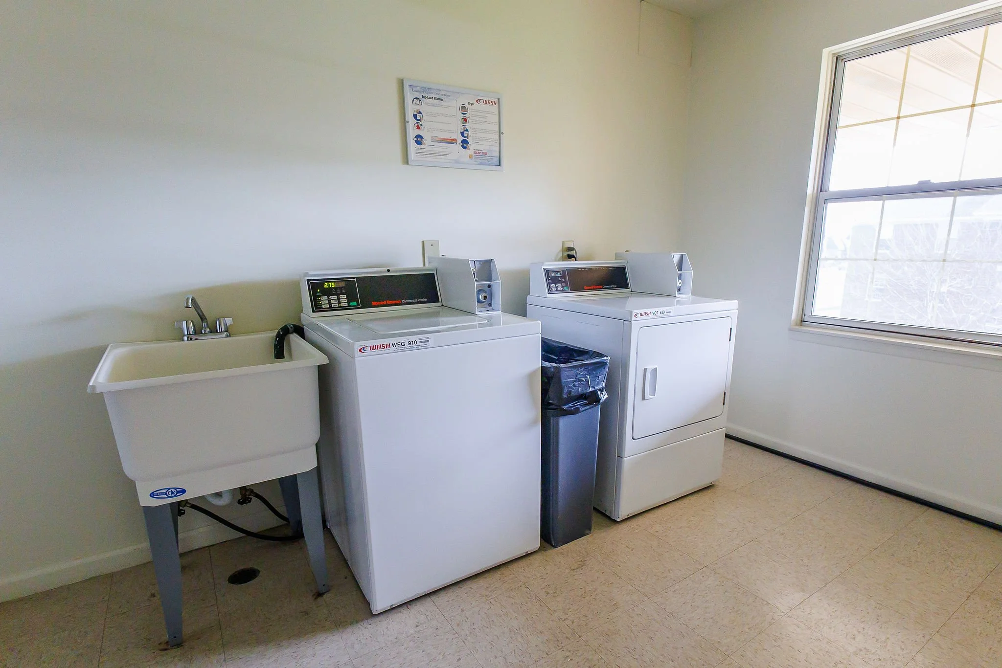 Laundry room with a utility sink, two washing machines, a trash can, a window, and a poster on the wall.