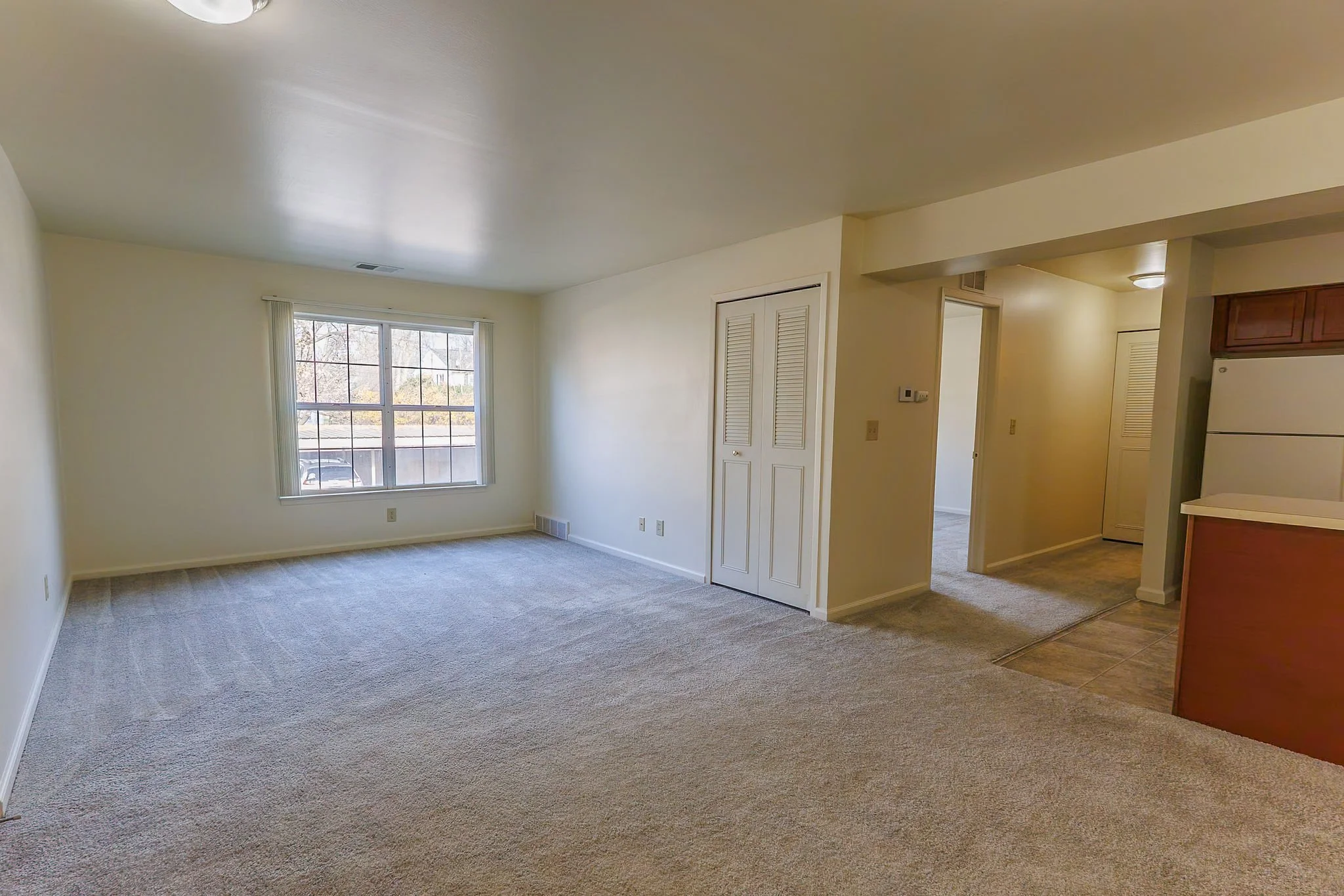 Empty living room with a large window, beige carpet, and beige painted walls. There is a white door with louvered panels, a thermostat on the wall, and a view into a hallway with a ceiling light. Part of a kitchen with wood cabinets and a white refrigerator is visible.