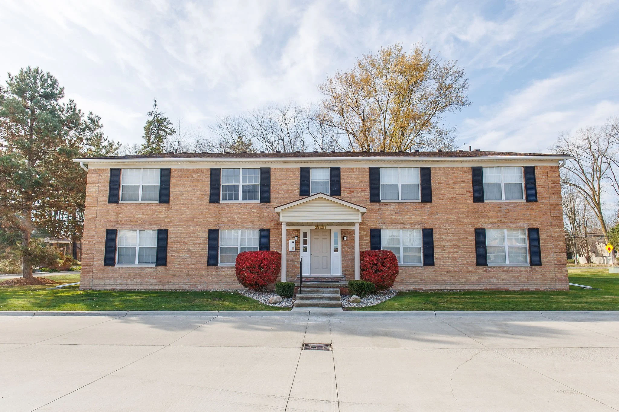 Two-story brick apartment building with black shutters, white windows, and a small entrance porch with steps, flanked by red bushes and green grass, under a partly cloudy sky.