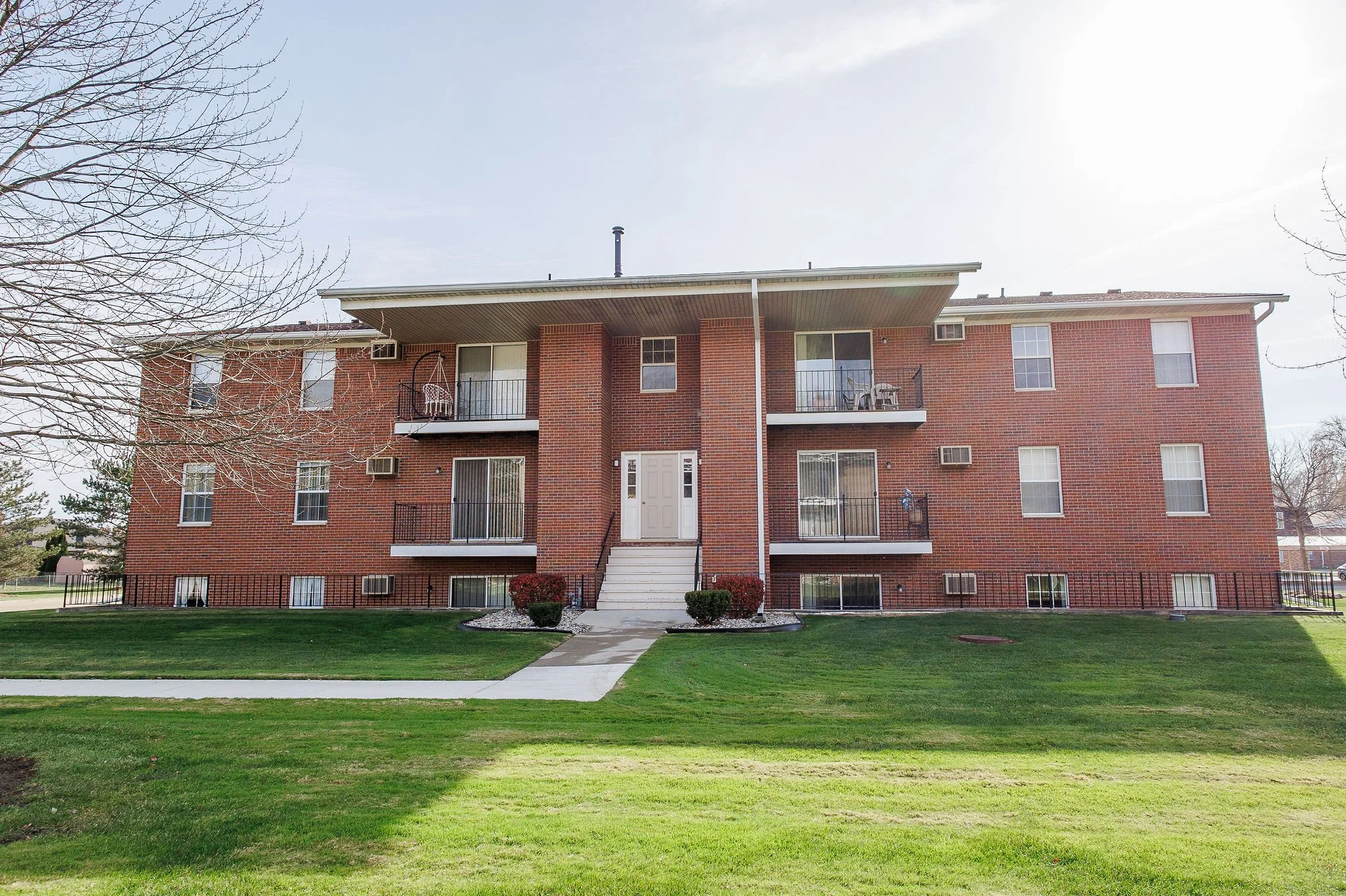 A three-story red brick apartment building with multiple balconies and windows, surrounded by well-maintained grass and landscaping in a suburban area.