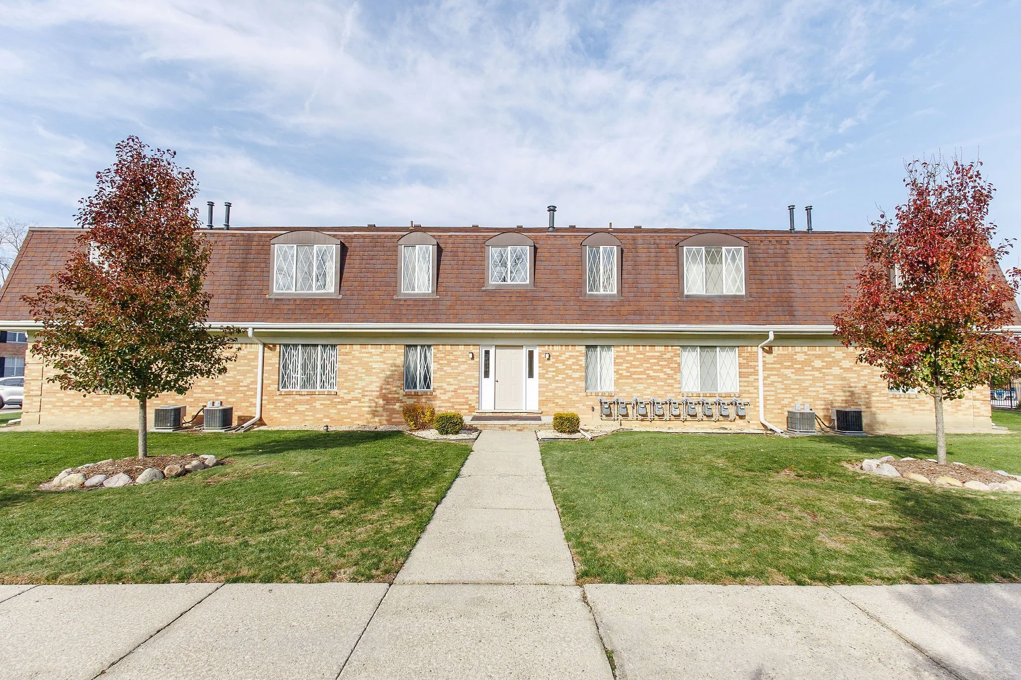 Two-story brick residential building with a brown shingle roof, seven windows with diamond-patterned trim, white front door, landscaped front yard with grass, two trees with red leaves, and a concrete walkway leading to the entrance, under a partly c