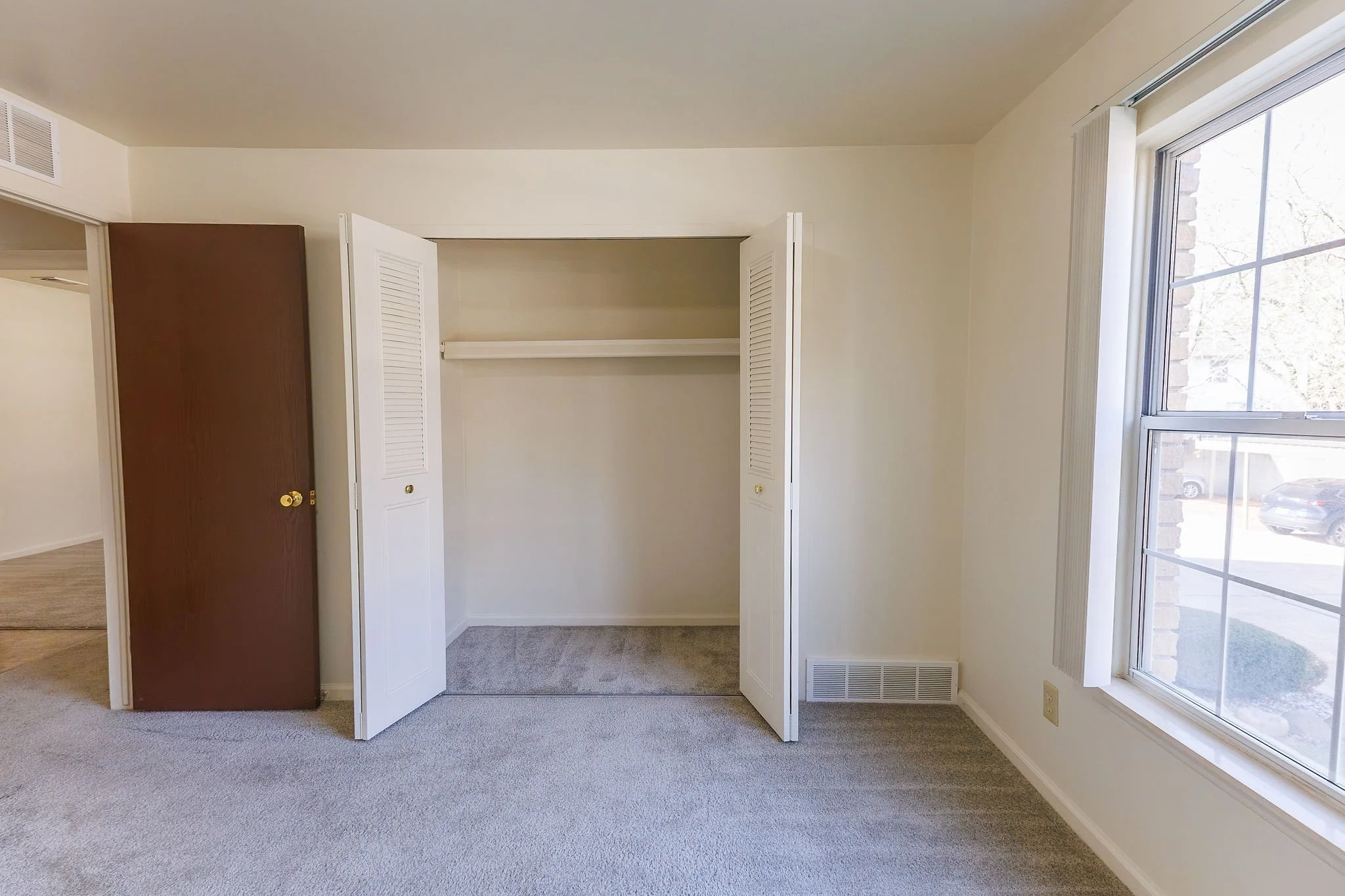 Empty room with a closet featuring bifold doors, a window with a view of parked cars outside, and beige carpeting.