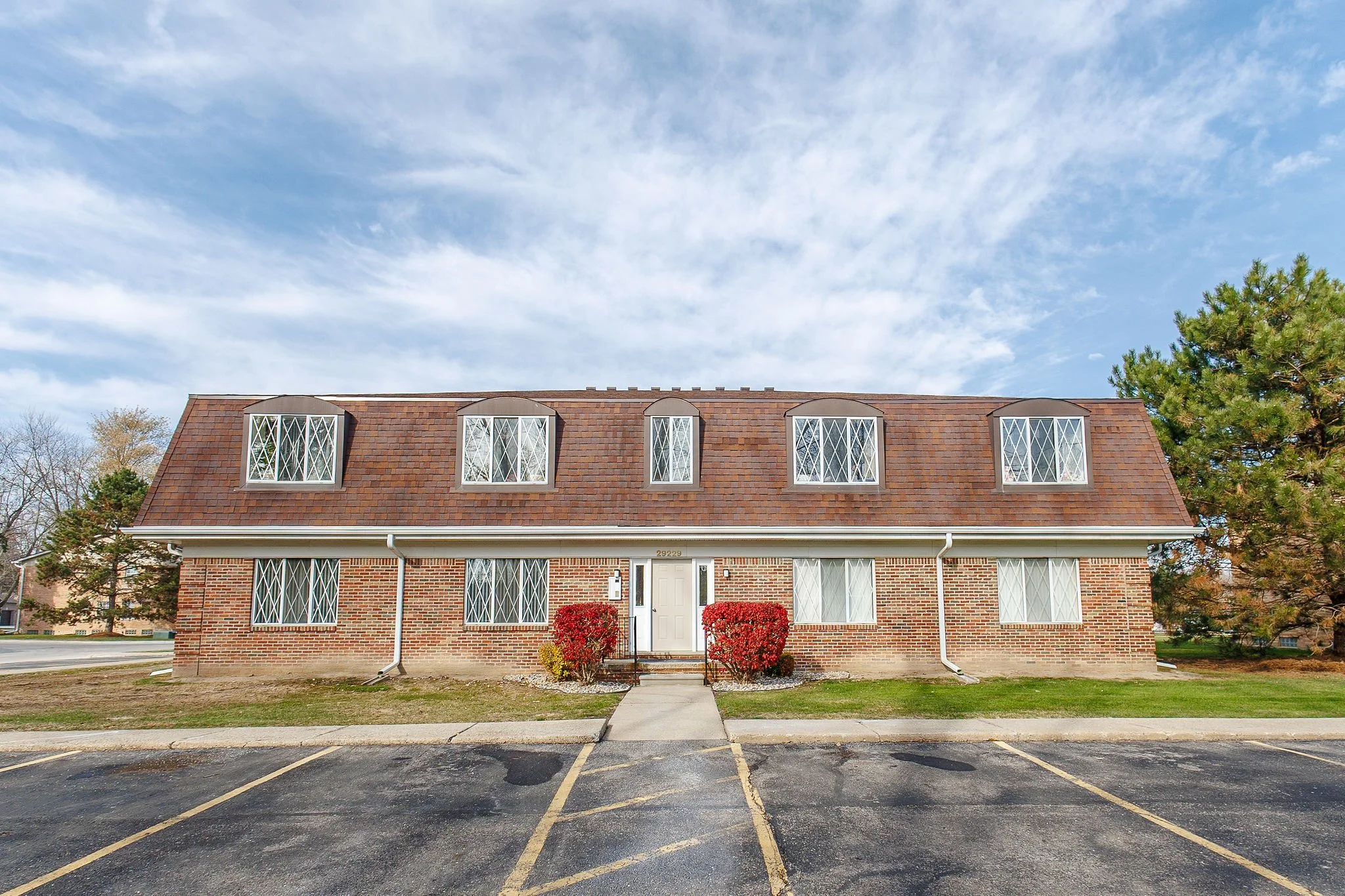 A two-story brick apartment building with a brown shingled roof, white framed windows with lattice design, and a white front door. There are two red bushes flanking the front steps, and a parking lot in front with yellow parking lines. The sky is par