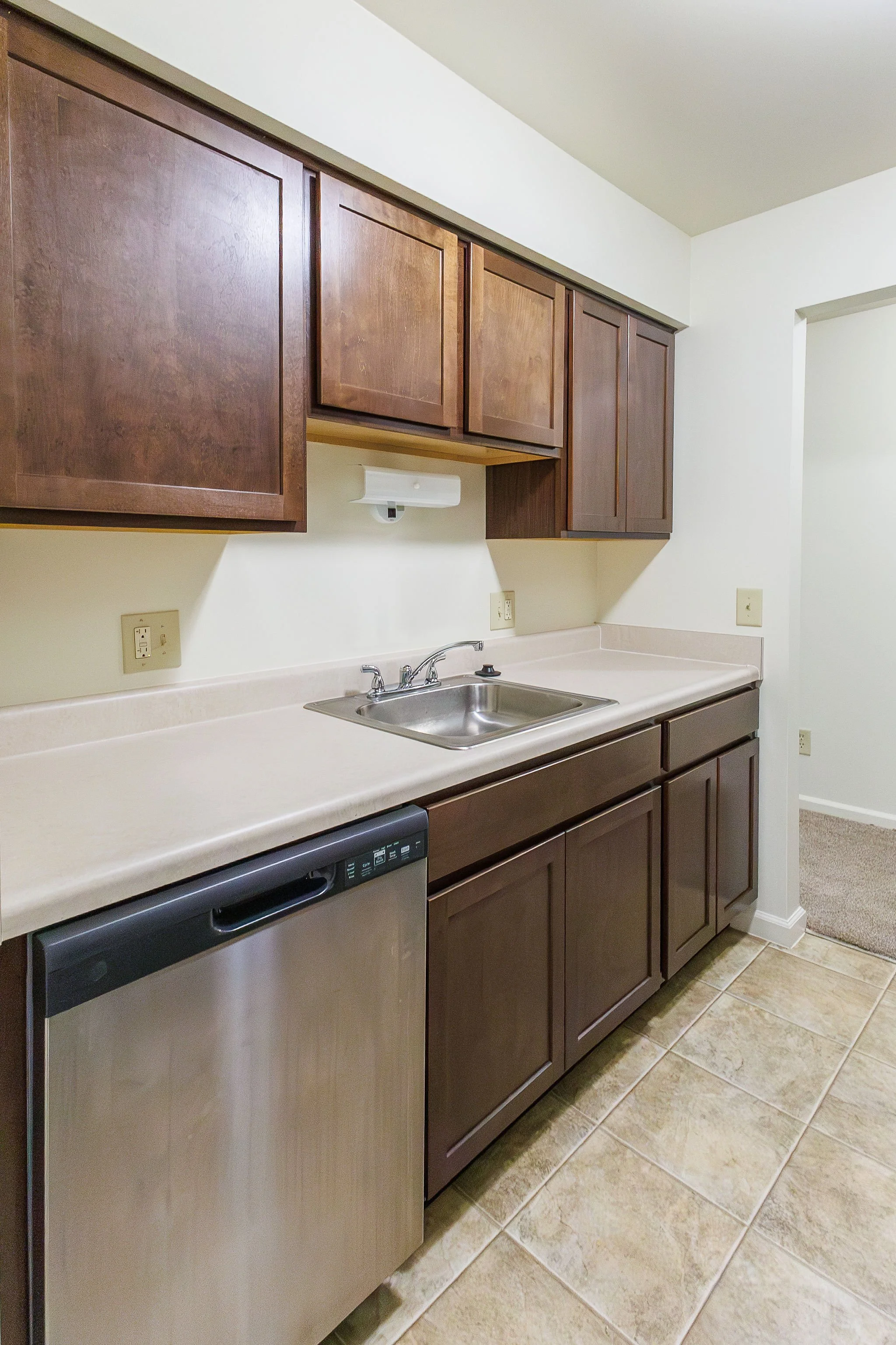 Kitchen with brown wooden cabinets, a stainless steel sink, beige countertop, and a dishwasher, with tiled floor.