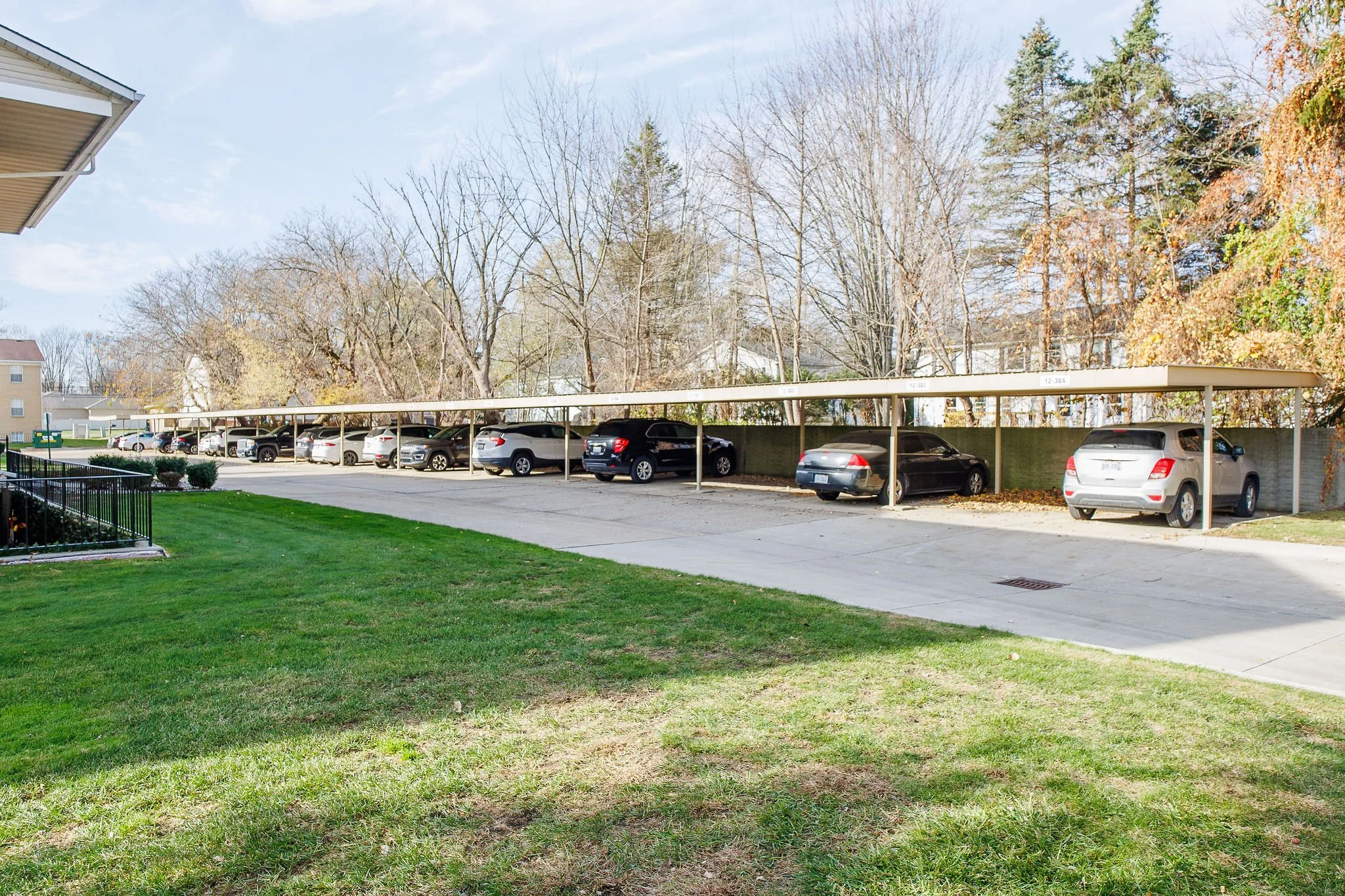 Parking lot with multiple parked cars under carport, adjacent to grassy area with trees in the background, residential buildings visible.