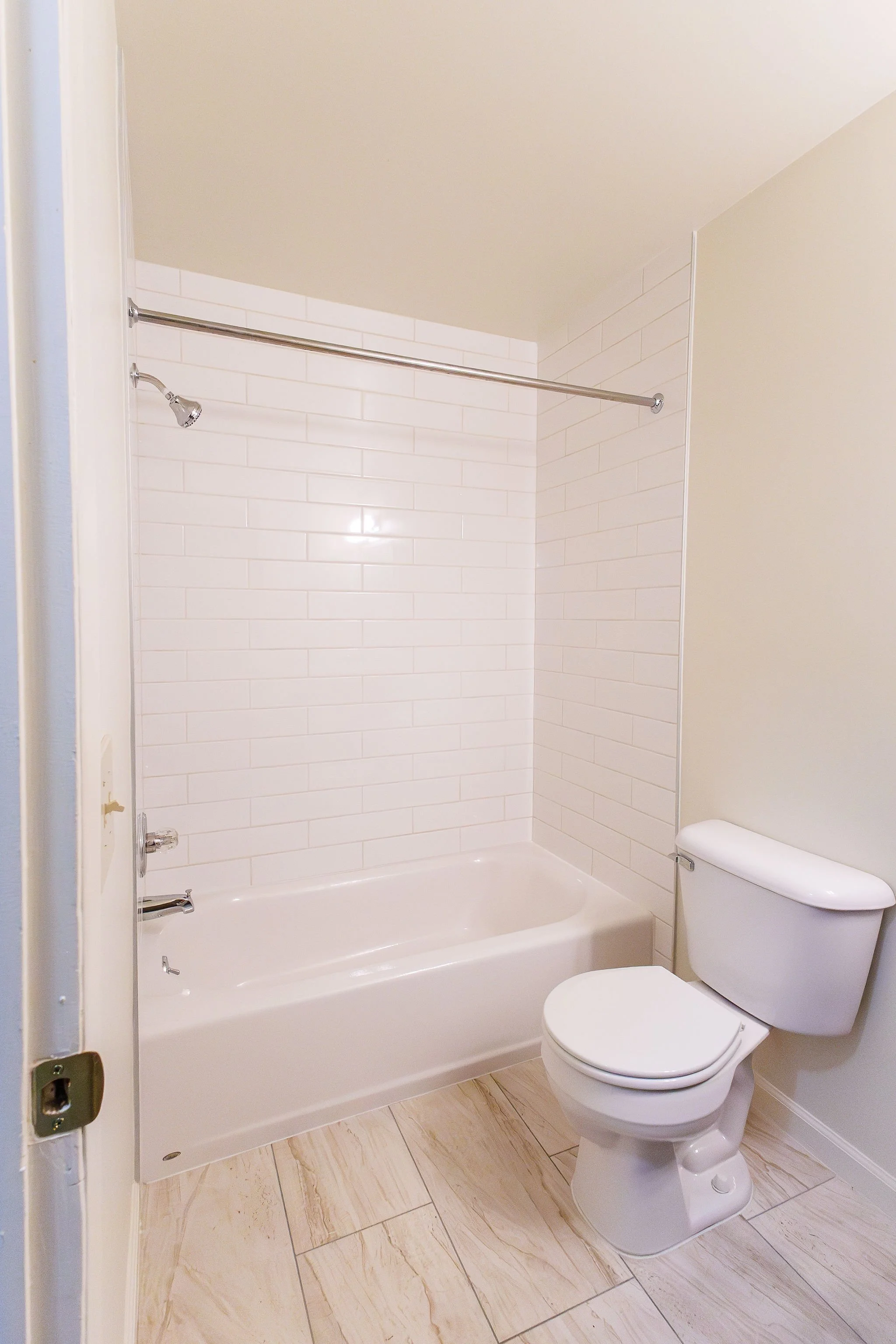 Bathroom with a bathtub, shower, and toilet, neutral-colored walls, white tiled wall in the shower area, and light-colored floor tiles.