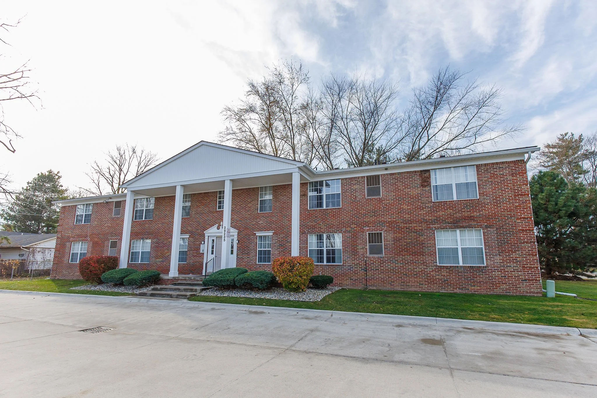 Two-story brick apartment building with white columns and entrance, surrounded by landscaped bushes and grass, under a partly cloudy sky.