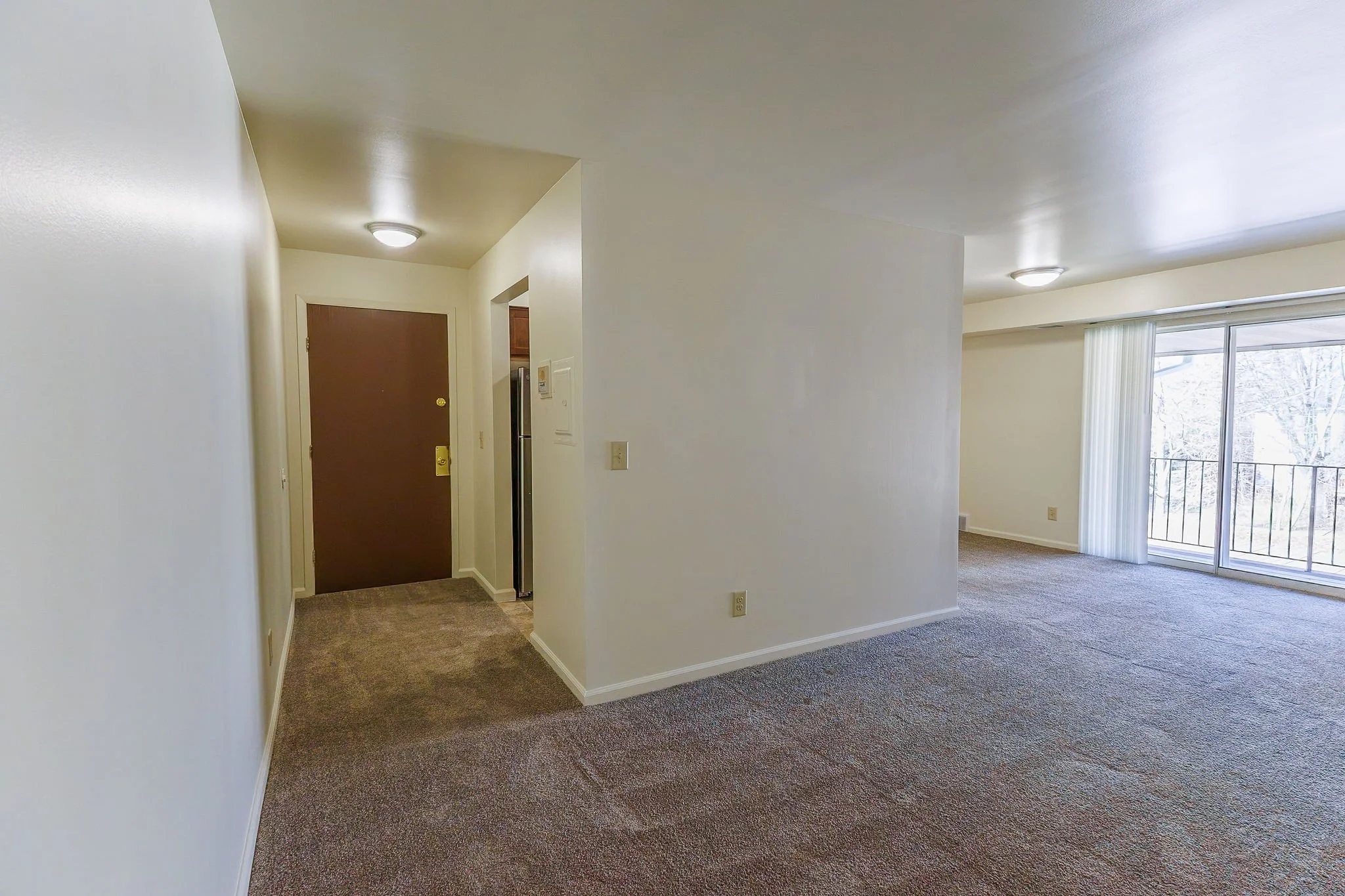 Empty living room with large window, sliding glass door, and balcony, beige carpet, white walls, and ceiling, with a brown front door and a small hallway.