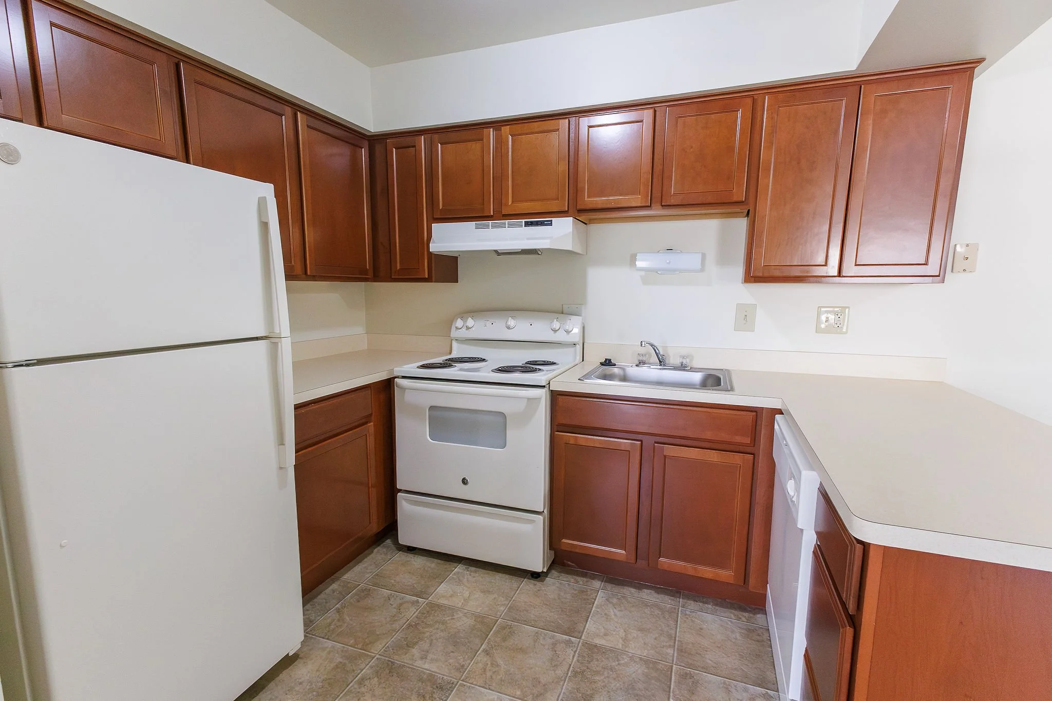 Kitchen with white refrigerator, white stove, and brown wood cabinets, beige countertops, and beige tiled floor.
