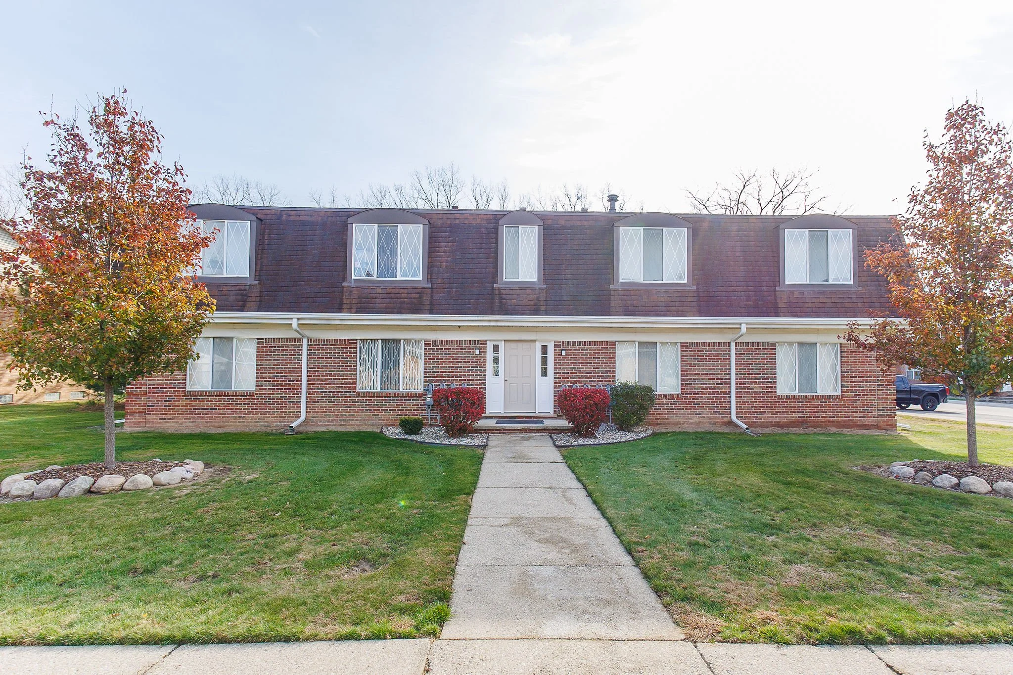 A two-story brick residential building with a pathway leading to the front door, surrounded by manicured lawns and trees with fall foliage.