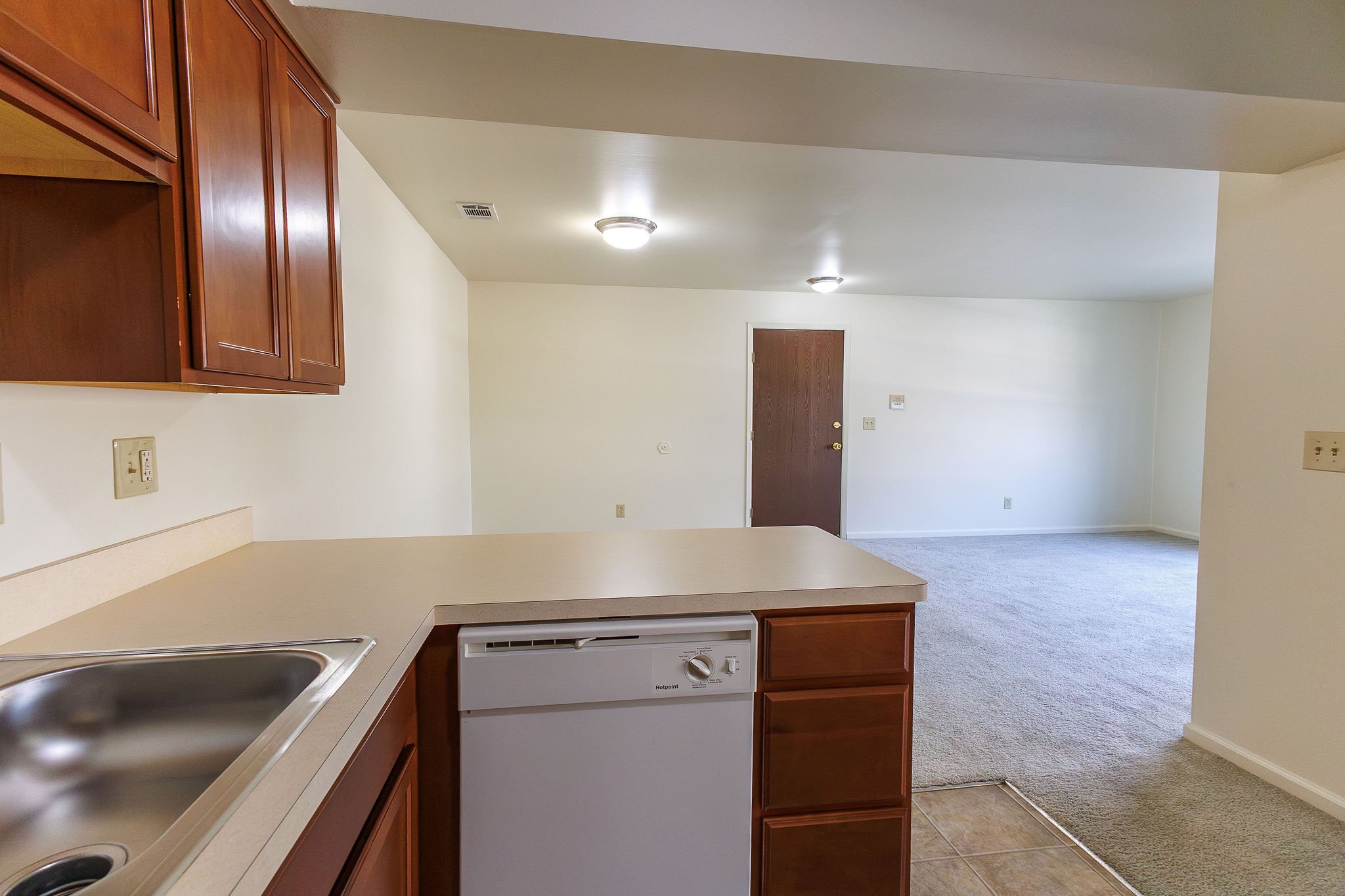 Empty living room and kitchen area of an apartment with a white wall, wooden cabinets, beige countertops, and a combination of carpet and tile flooring.
