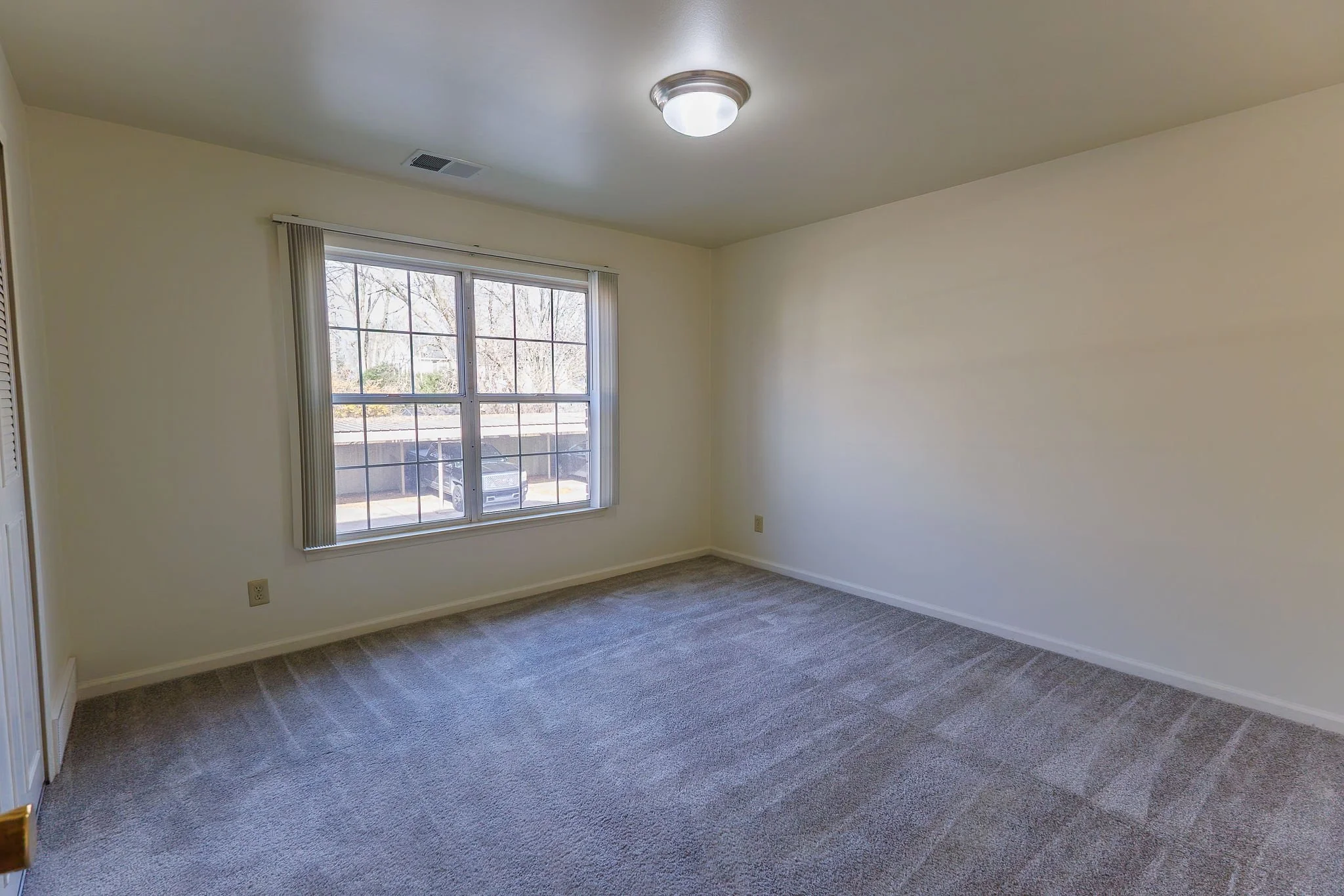 Empty bedroom with beige walls, gray carpet, a large window, and a ceiling light.