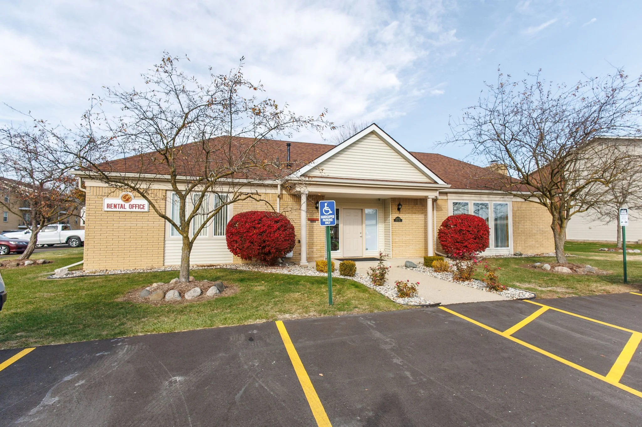 A single-story building with a beige brick exterior, white door, and large windows. There are two leafless trees and two red bushes in front. A parking lot with yellow lines is in the foreground. A sign indicates a handicap parking space, and a sign on the building reads "Rental Office."