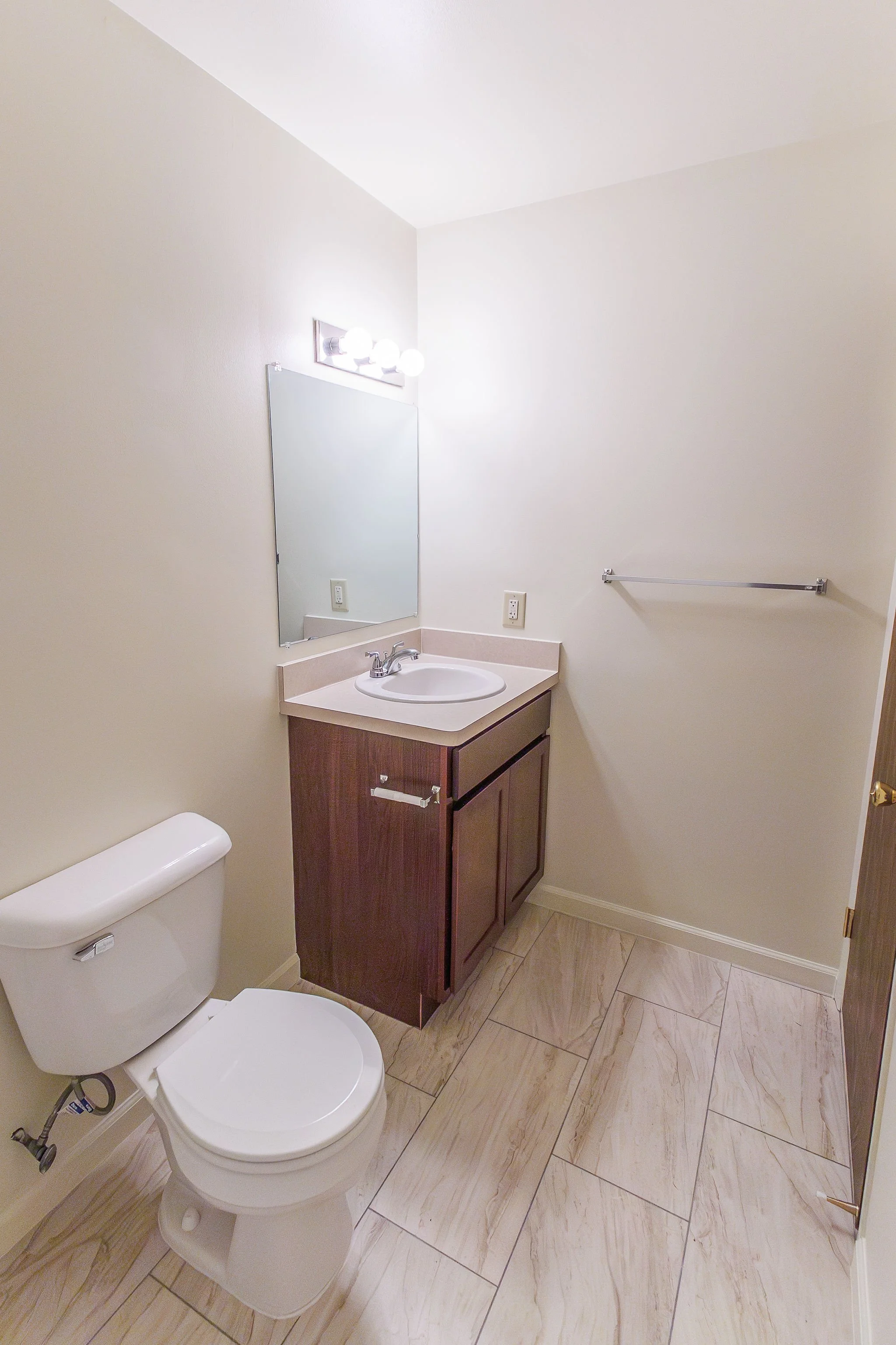 Small bathroom with a white toilet, a wooden vanity with a white sink, a mirror, a towel bar, and beige-tiled flooring.