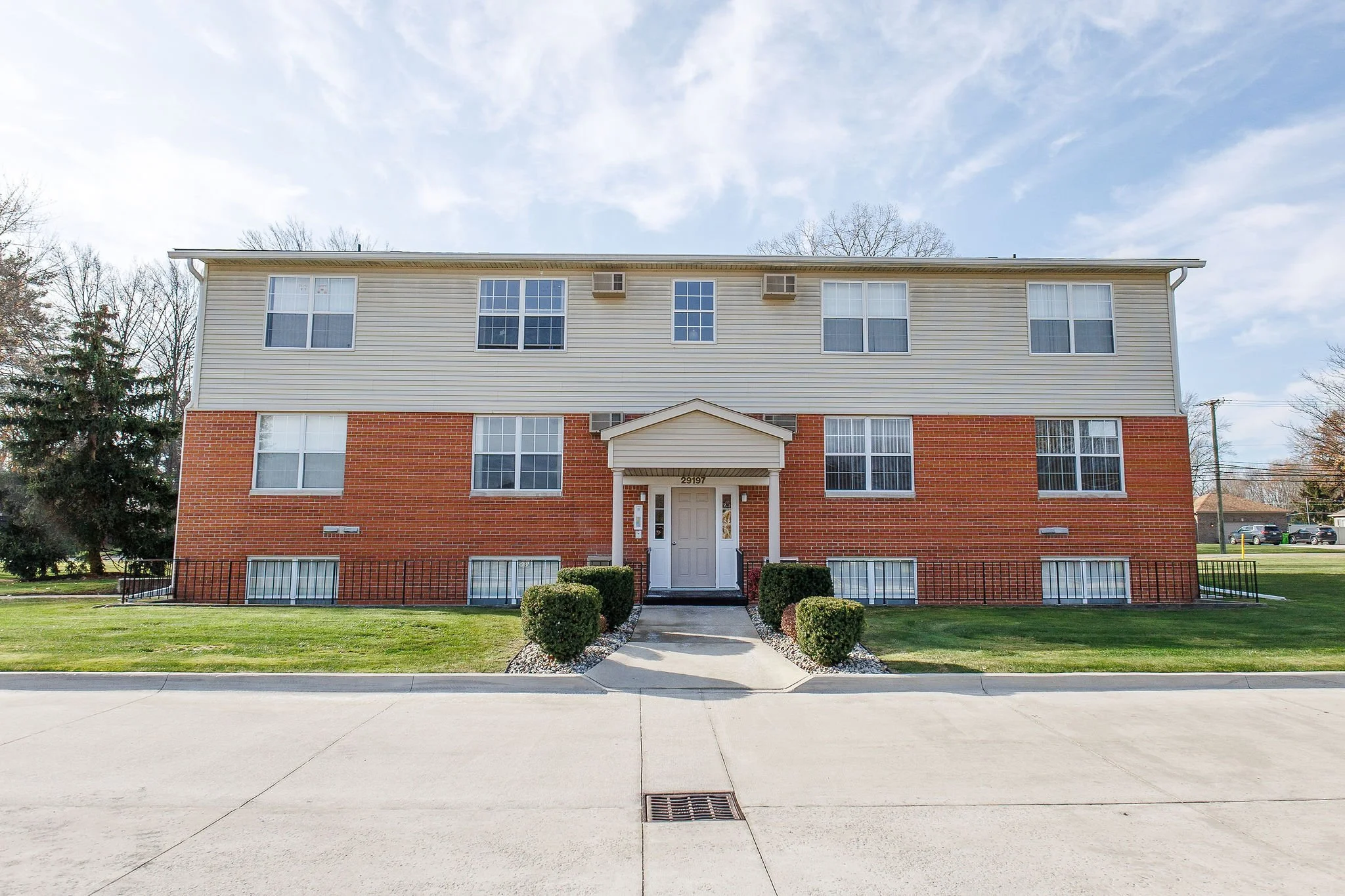 A three-story apartment building with a brick lower level and beige vinyl siding upper level. It has a centered front door with a small gabled porch, surrounded by neatly trimmed bushes and a sidewalk, with a grassy lawn and trees in the background.