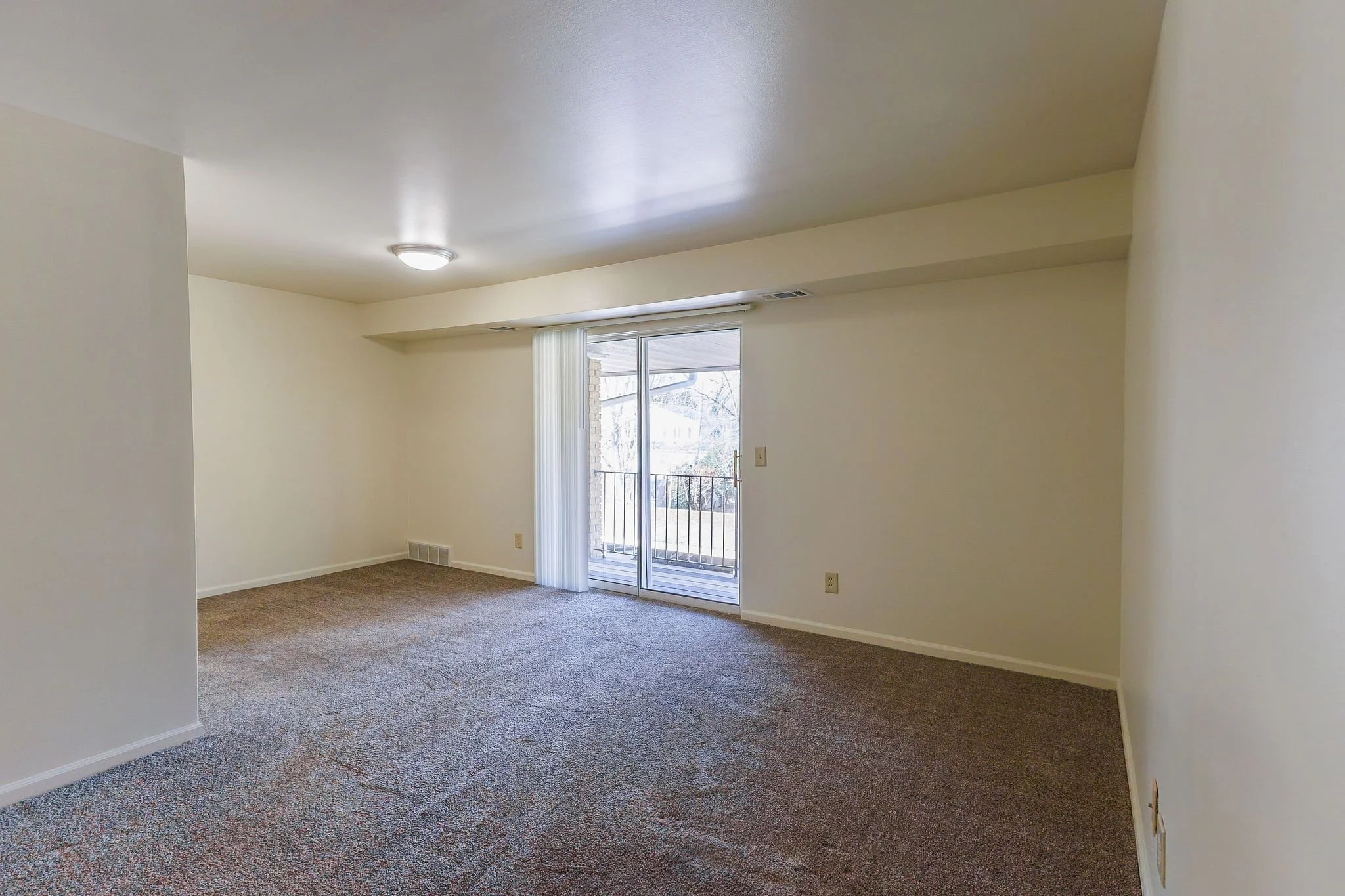 Empty living room with sliding glass door leading to a balcony, neutral-colored walls, brown carpet flooring, and ceiling light fixture.