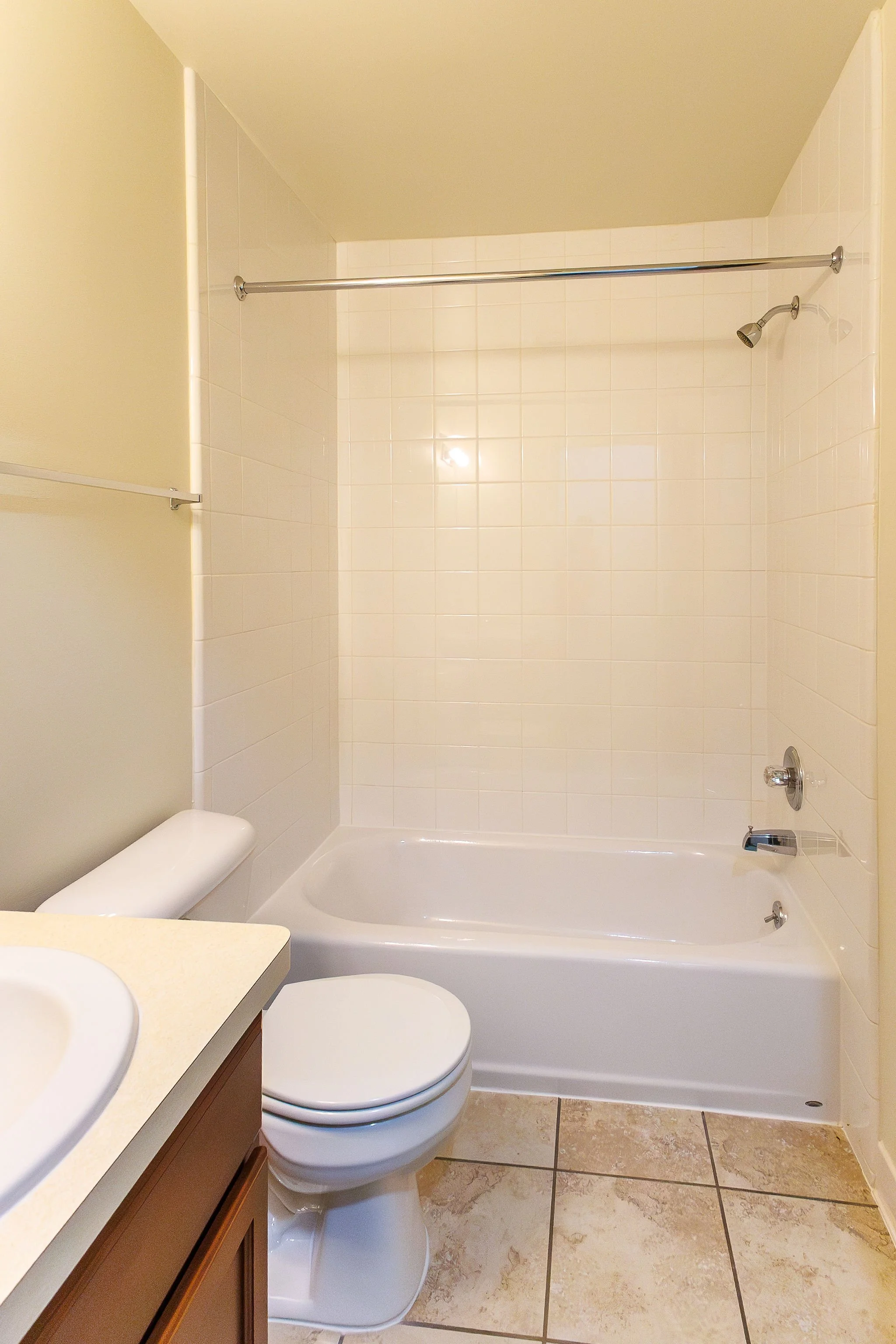 A bathroom with a bathtub, a showerhead, a toilet, and a sink with a brown cabinet. The walls are tiled in white, and the floor has beige tiles. There is a curved shower curtain rod installed above the bathtub.