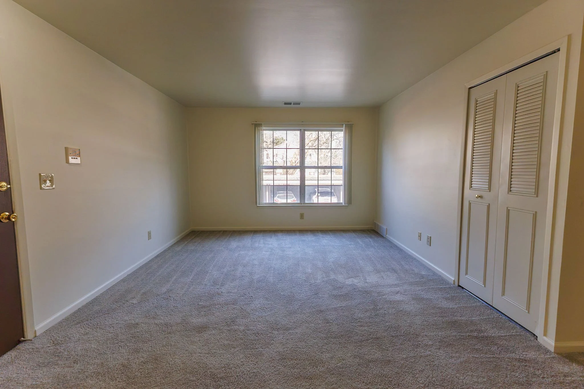 Empty living room with beige carpet, white walls, a window with blinds, and a closed closet door.