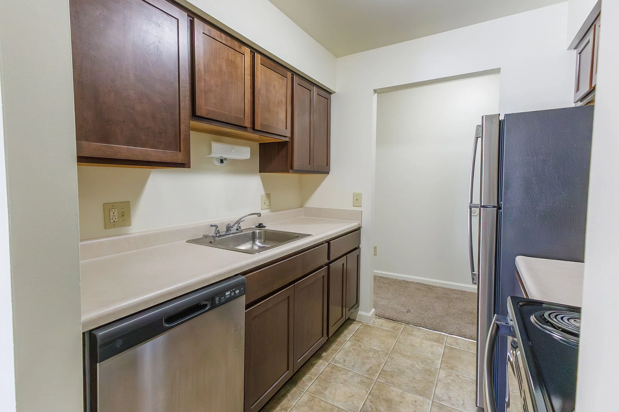 A small kitchen with beige countertops, oak cabinets, a stainless steel sink, a dishwasher, a refrigerator, and a stove. The floor is tiled in beige, and there is a small carpeted area at the doorway.