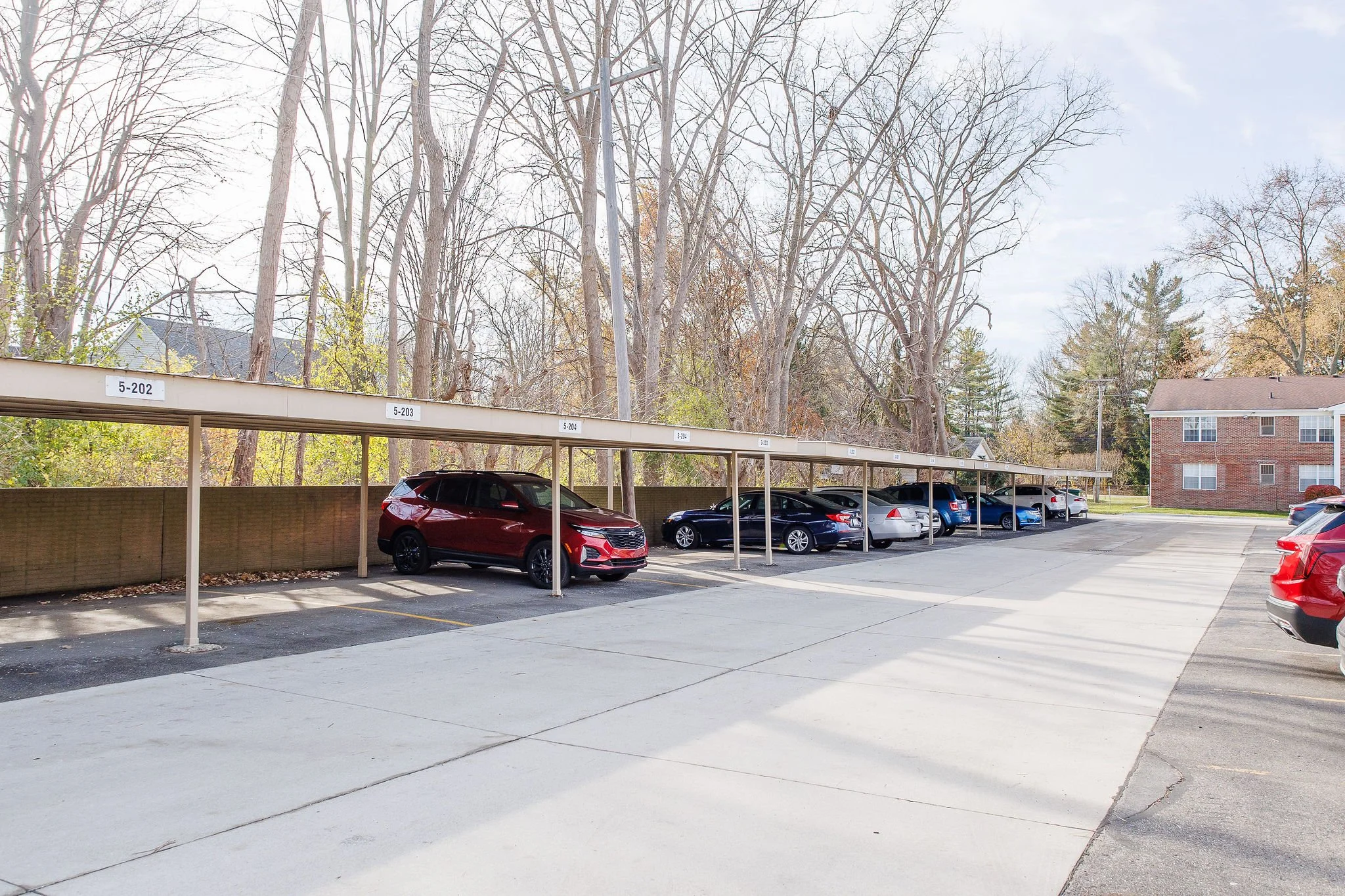 Empty outdoor parking lot with parked cars along a covered parking structure, leafless trees in the background, and a brick building to the right