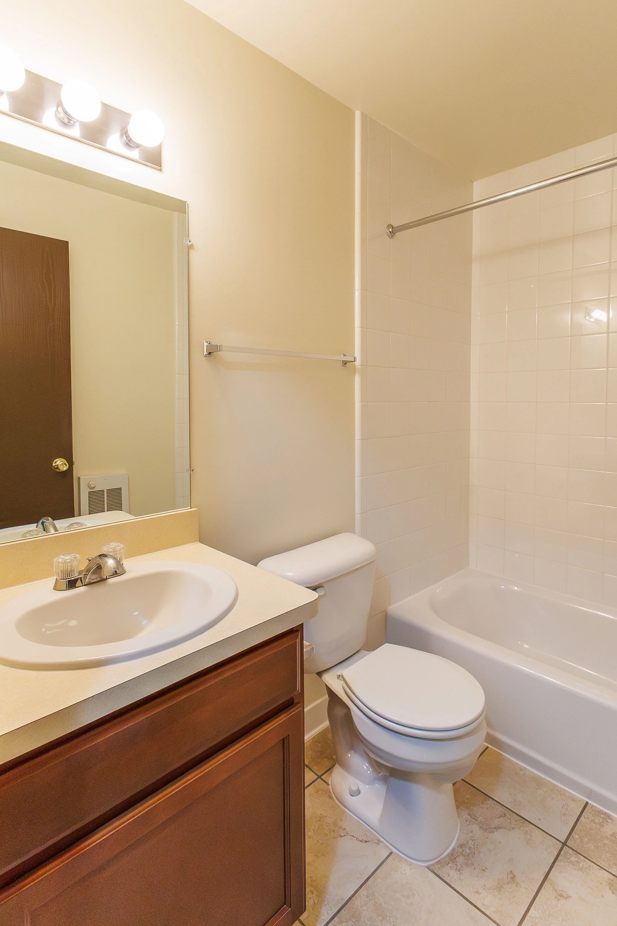 A small bathroom featuring a sink with a mirror above, a toilet, and a bathtub with a shower curtain rod. The room has beige tiled flooring and light-colored walls.