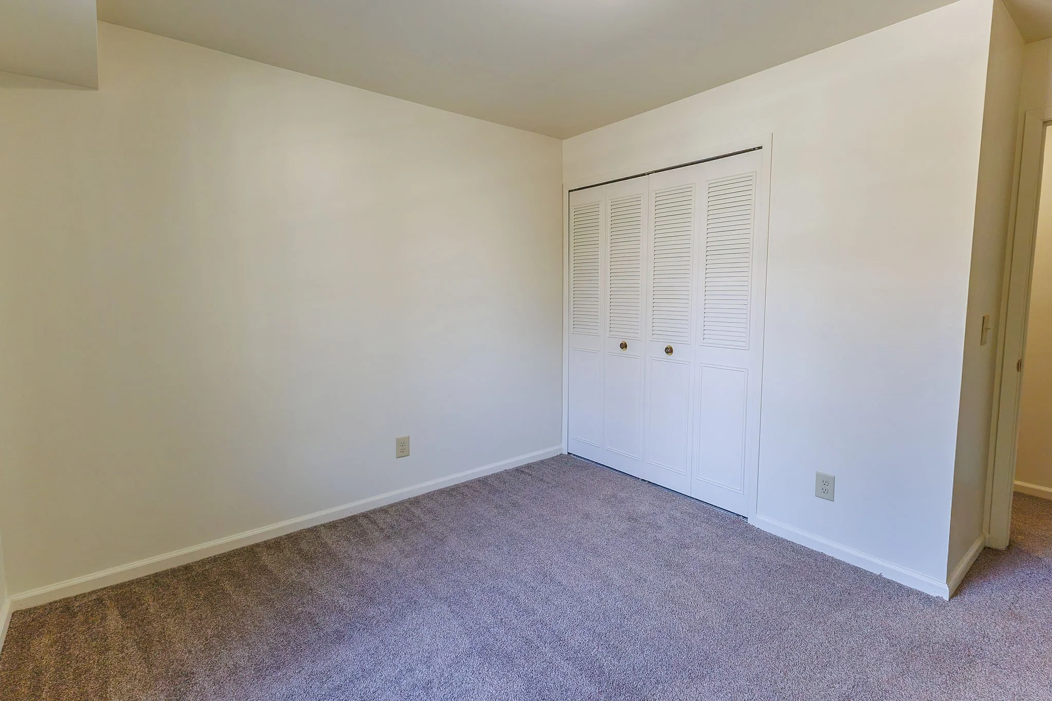 Empty room with white walls, closed closet doors, carpeted floor, and electrical outlets.
