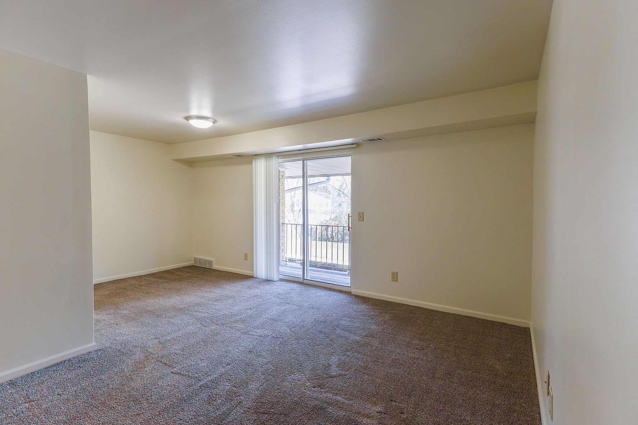 Empty living room with sliding glass door leading to balcony, white walls, beige carpet, and ceiling light.