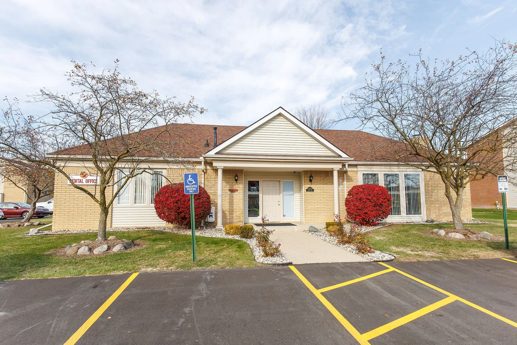 A single-story beige brick building with a gabled roof, white columns, and an entrance door, surrounded by trees and landscaped with red bushes and rocks; a parking lot with yellow striped spaces is in the foreground.