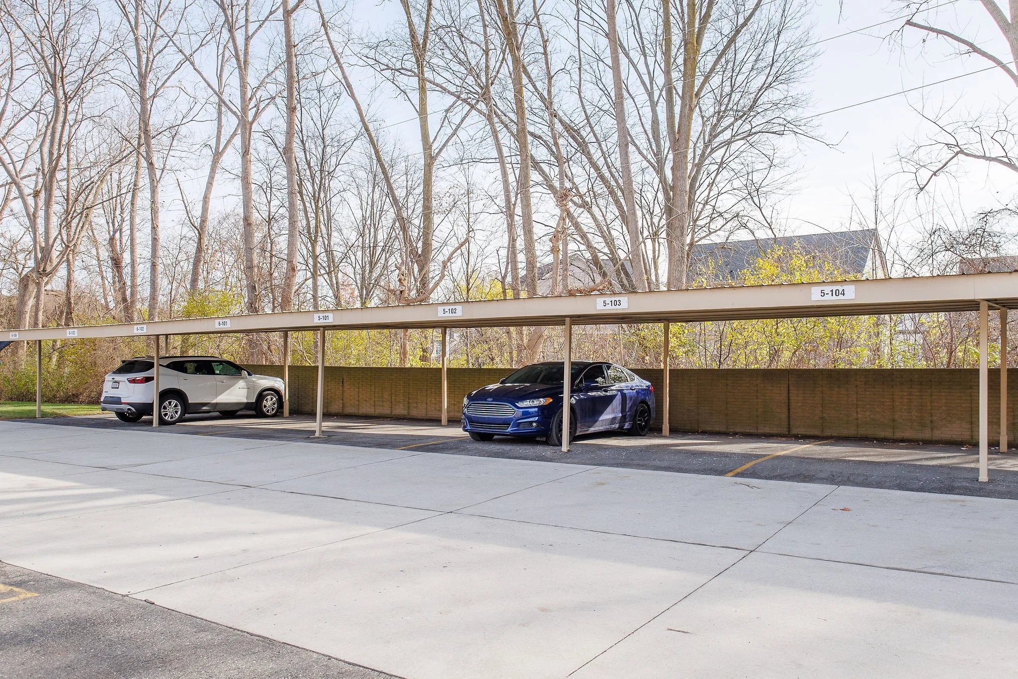 A parking lot with two parked cars under a beige carport with numbered signs, behind which there is a line of leafless trees and a building partially visible.