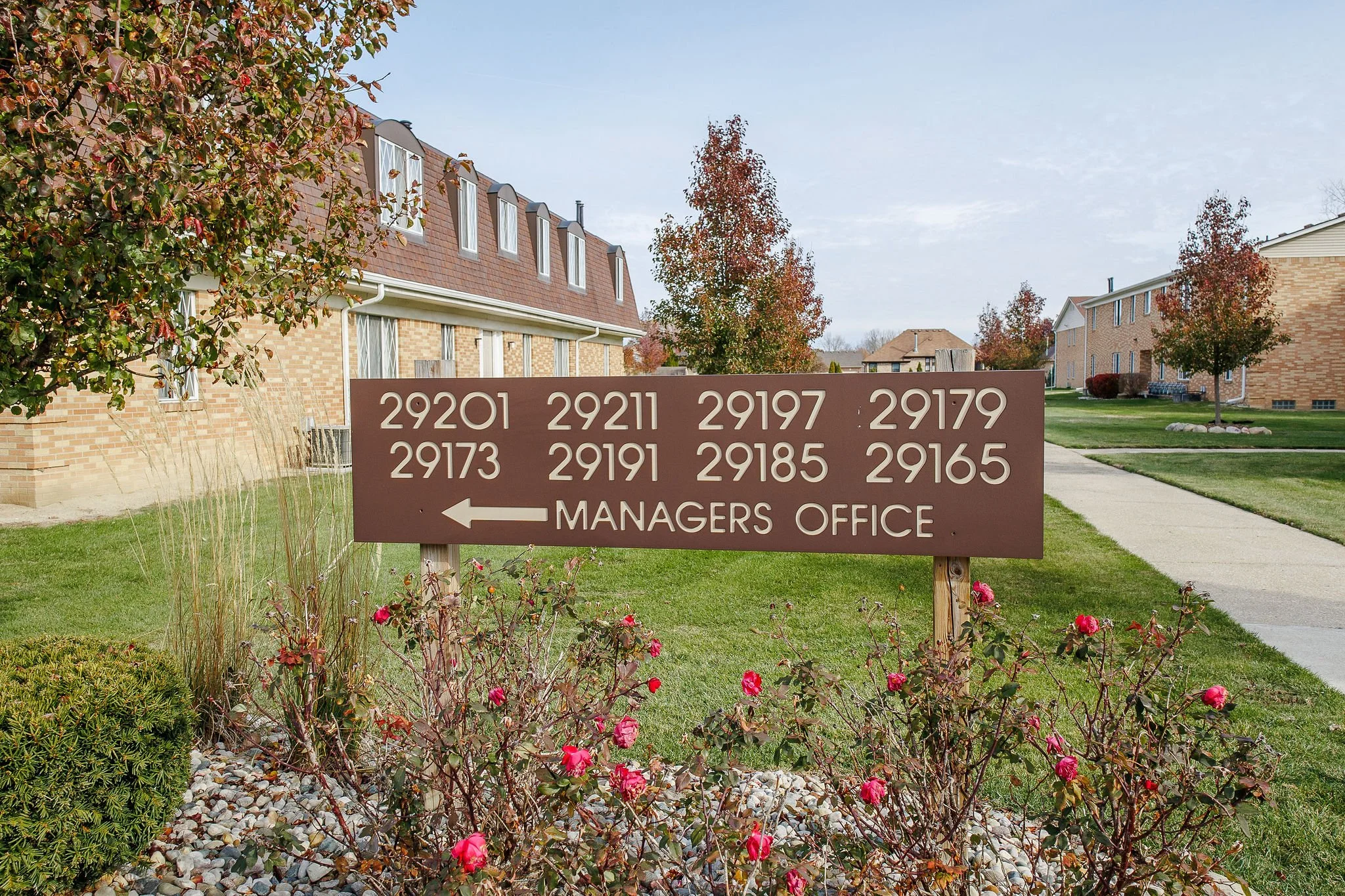 An outdoor sign with unit numbers and an arrow pointing left, indicating the direction to the managers office, in a residential complex with brick buildings, trees, and well-maintained grass.
