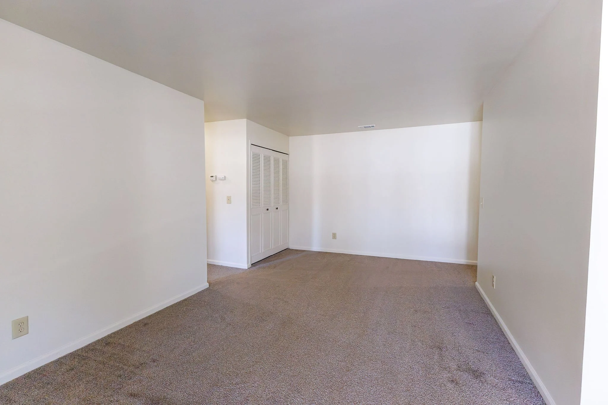 Empty living room with beige carpet, white walls, and a white ceiling; a closet with louvered doors and thermostat on the wall are visible.