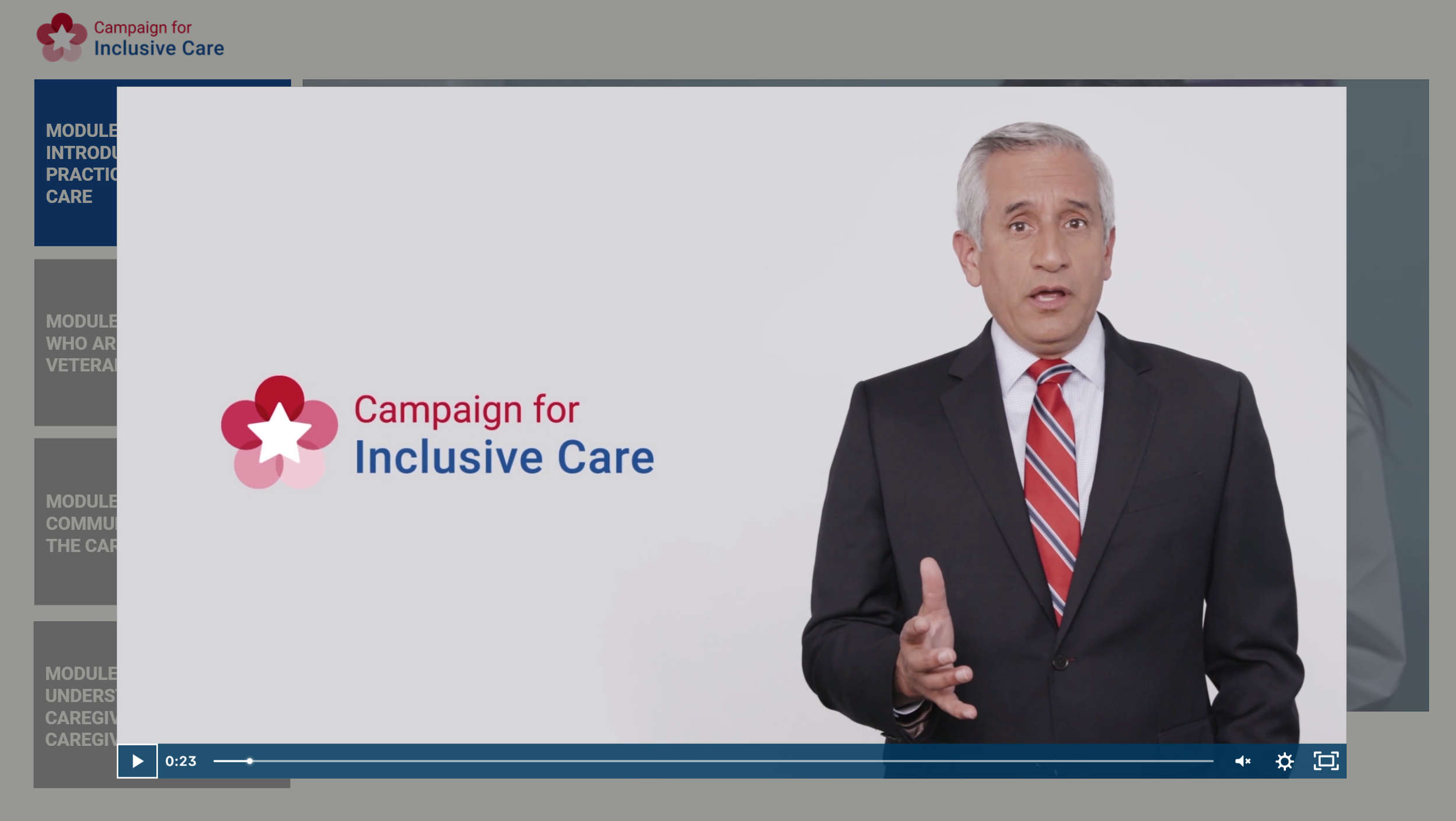 A man wearing a black suit, white shirt, and red striped tie standing in front of a white background with a slide titled 'Campaign for Inclusive Care' with a pink and red flower graphic and a white star in the center.