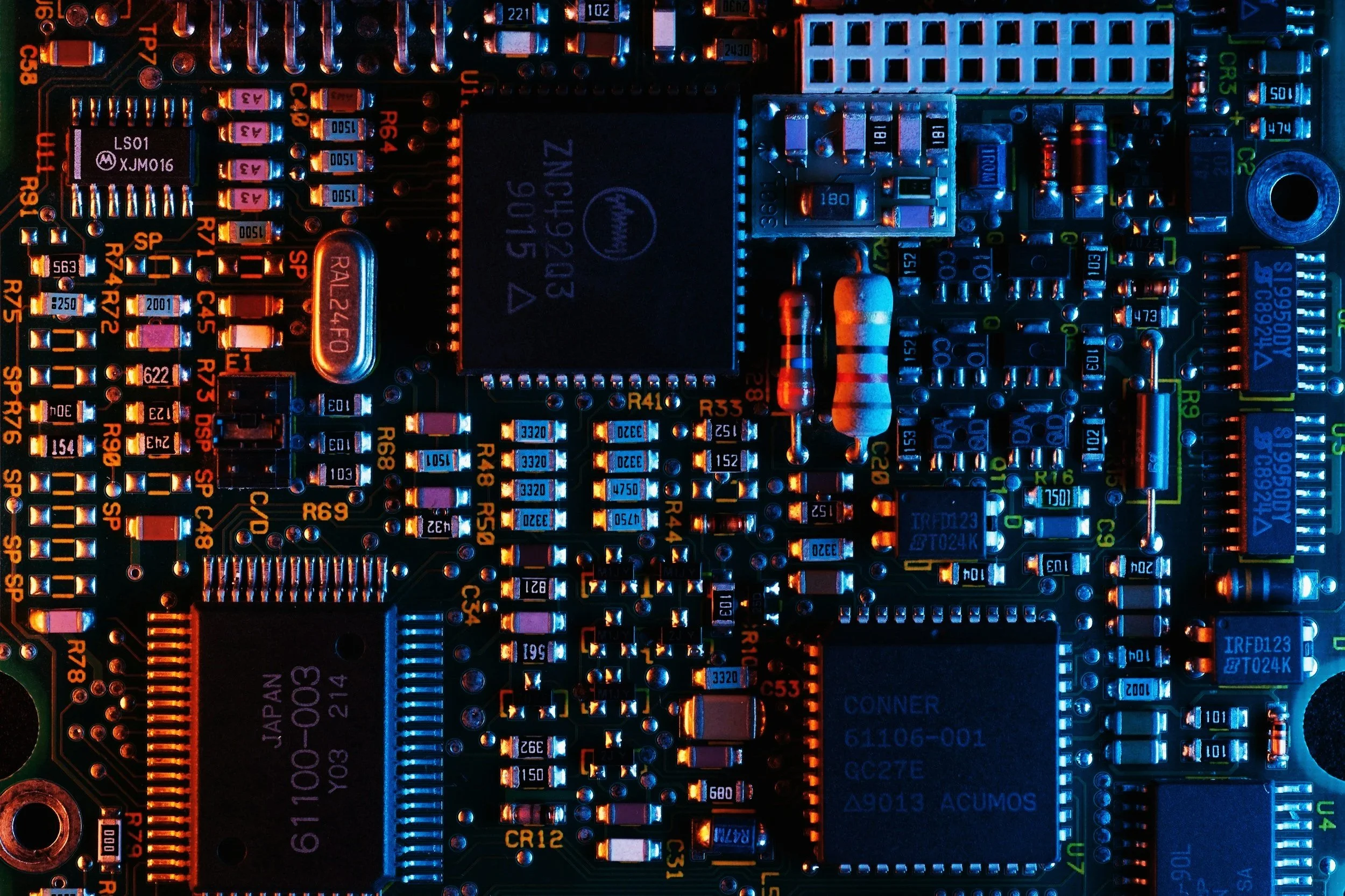 Placa de circuito eletrônico com componentes diversos, incluindo chips, resistores e capacitores, sob iluminação azul e laranja.