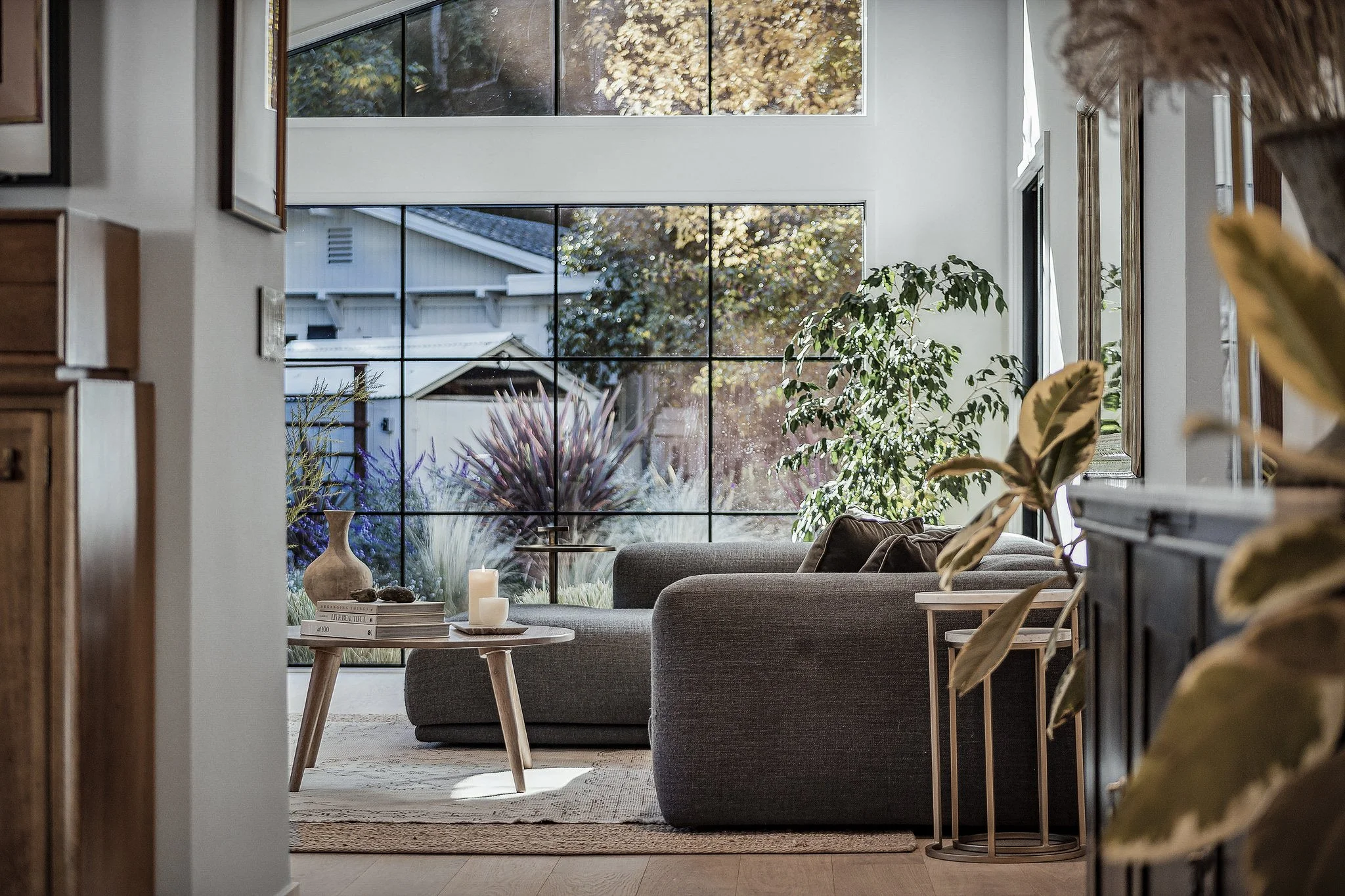 Living room with large window, gray sofa, wooden coffee table with candles, houseplants, and outdoor garden view.