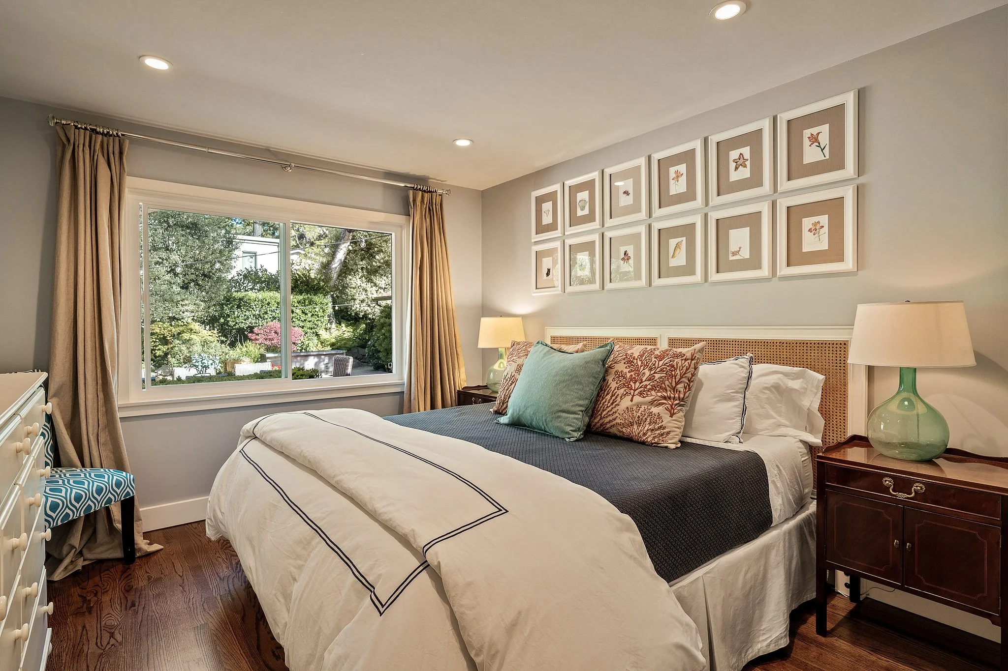 A bedroom with a large window, bed with multiple pillows, nightstand with lamp, and framed botanical prints on the wall.