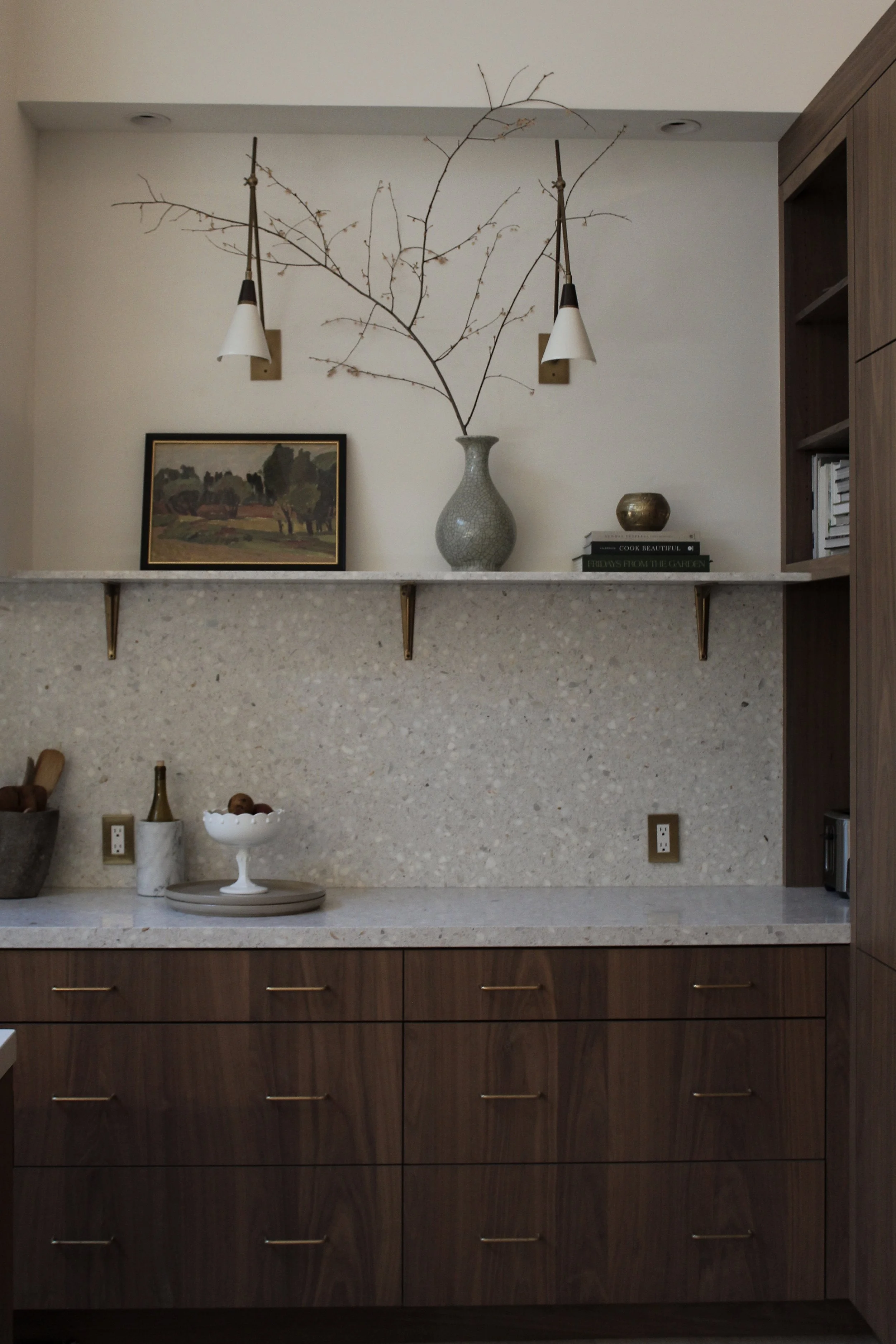 Kitchen backsplash with a countertop, decorative items, and a large vase with branches behind it, along with framed artwork, books, and small decorative objects on the shelf above.