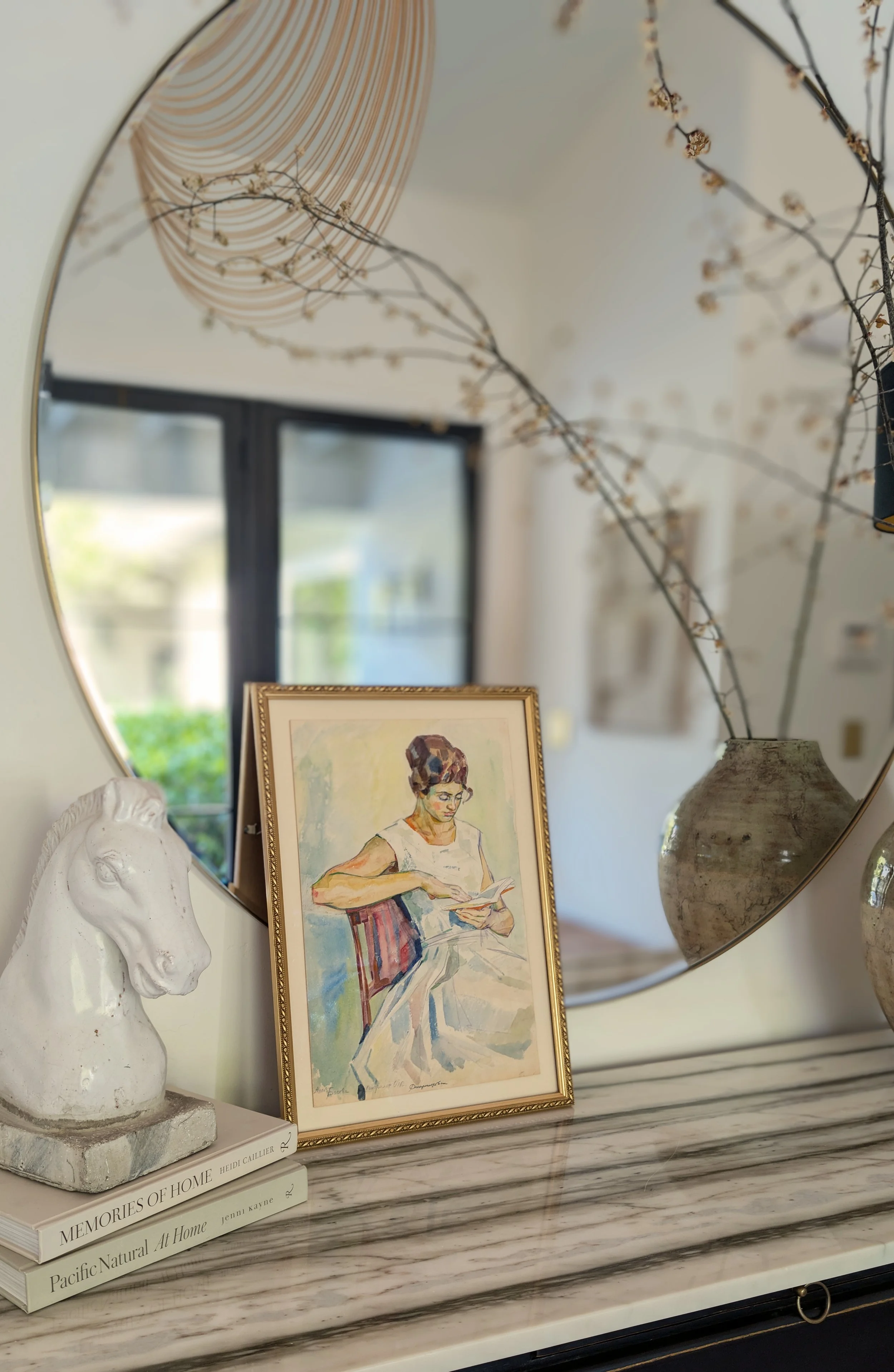 A marble tabletop with a decorative white horse bust, three stacked books, a framed painting of a woman reading, and two vases with dried branches, reflected in a round mirror.