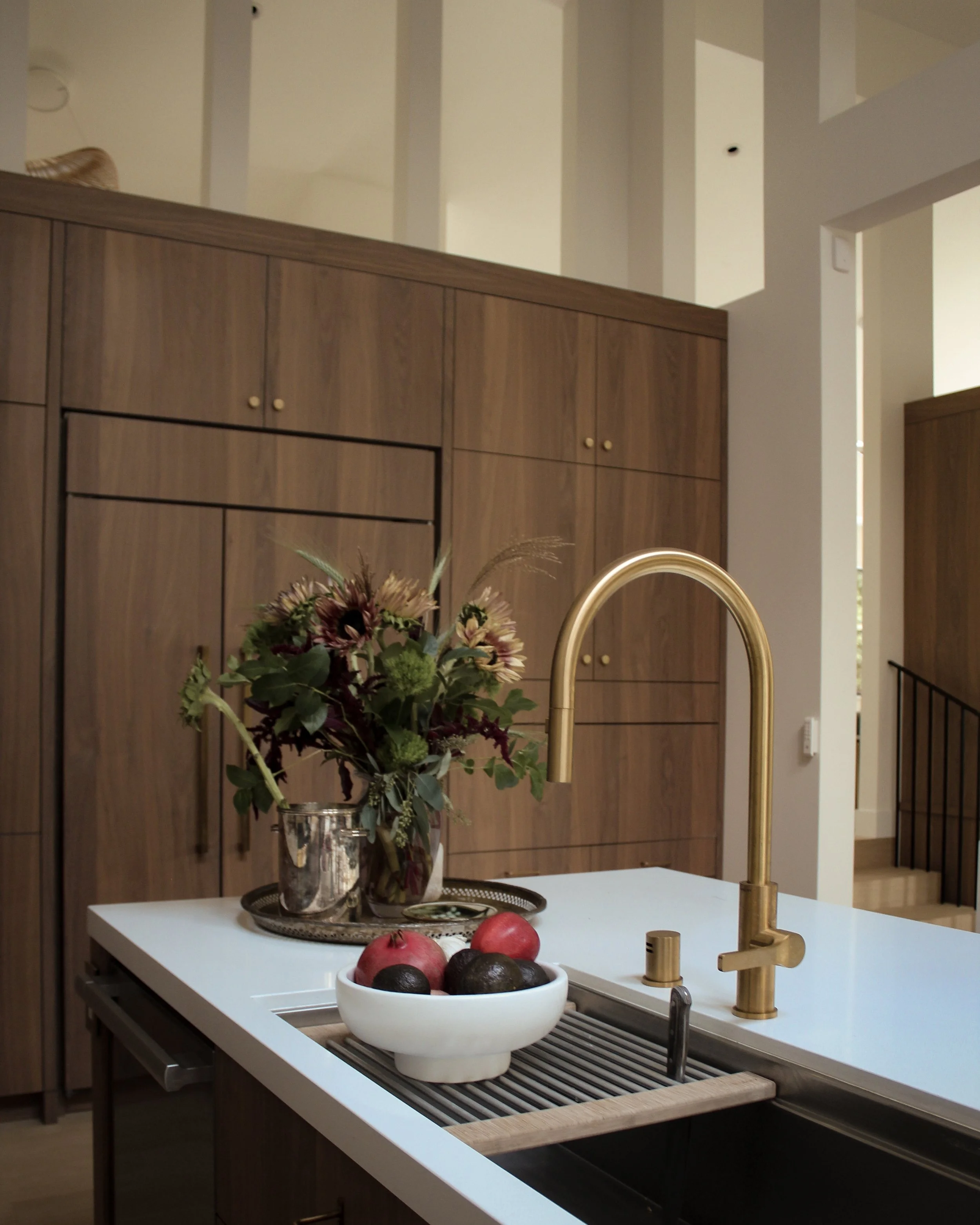 A modern kitchen island with a white countertop, a gold faucet, a bowl of fruits including pomegranates and avocados, and a bouquet of flowers in a silver vase. In the background, there are wooden cabinets with small round knobs.