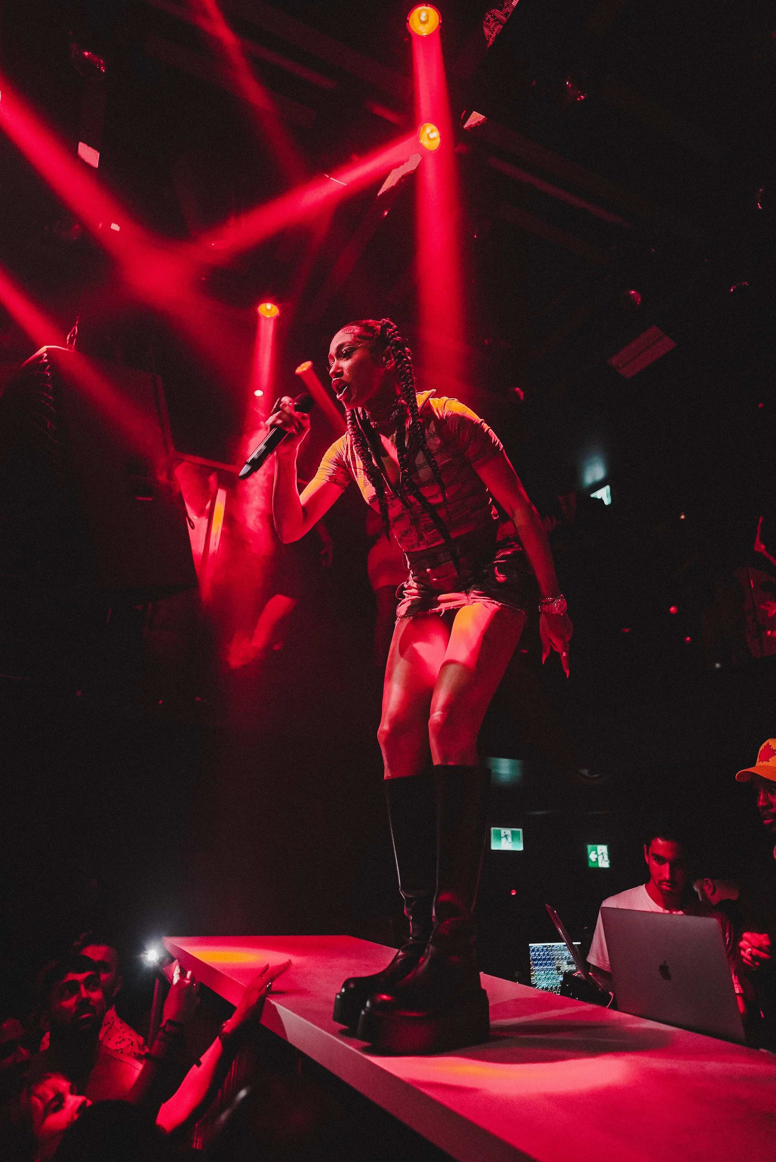 A female singer performs on stage with red lighting, holding a microphone, wearing a yellow top and black shorts. Audience members reach out towards her, and a sound technician is working on a laptop nearby.