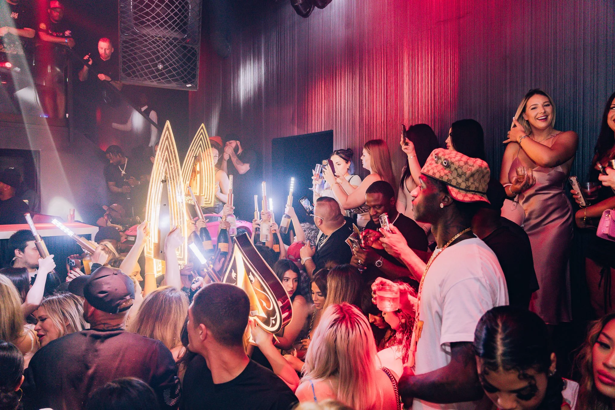 A crowded nightclub scene with people dancing, drinking, and socializing under red and purple lighting, with a DJ booth and neon lights.