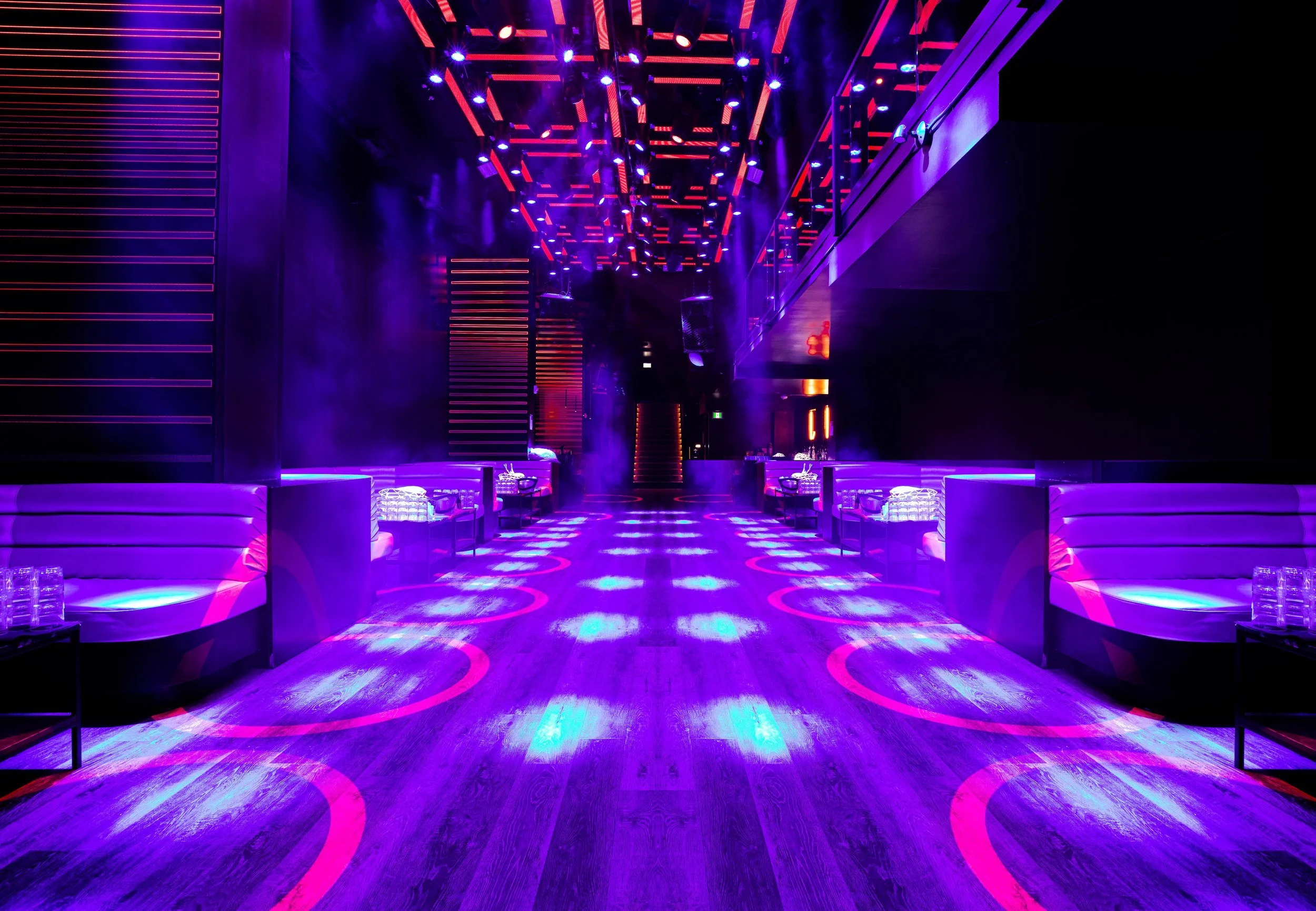 A futuristic nightclub interior with purple and pink neon lighting, curved seating, and a dance floor illuminated by circular purple and pink light patterns.