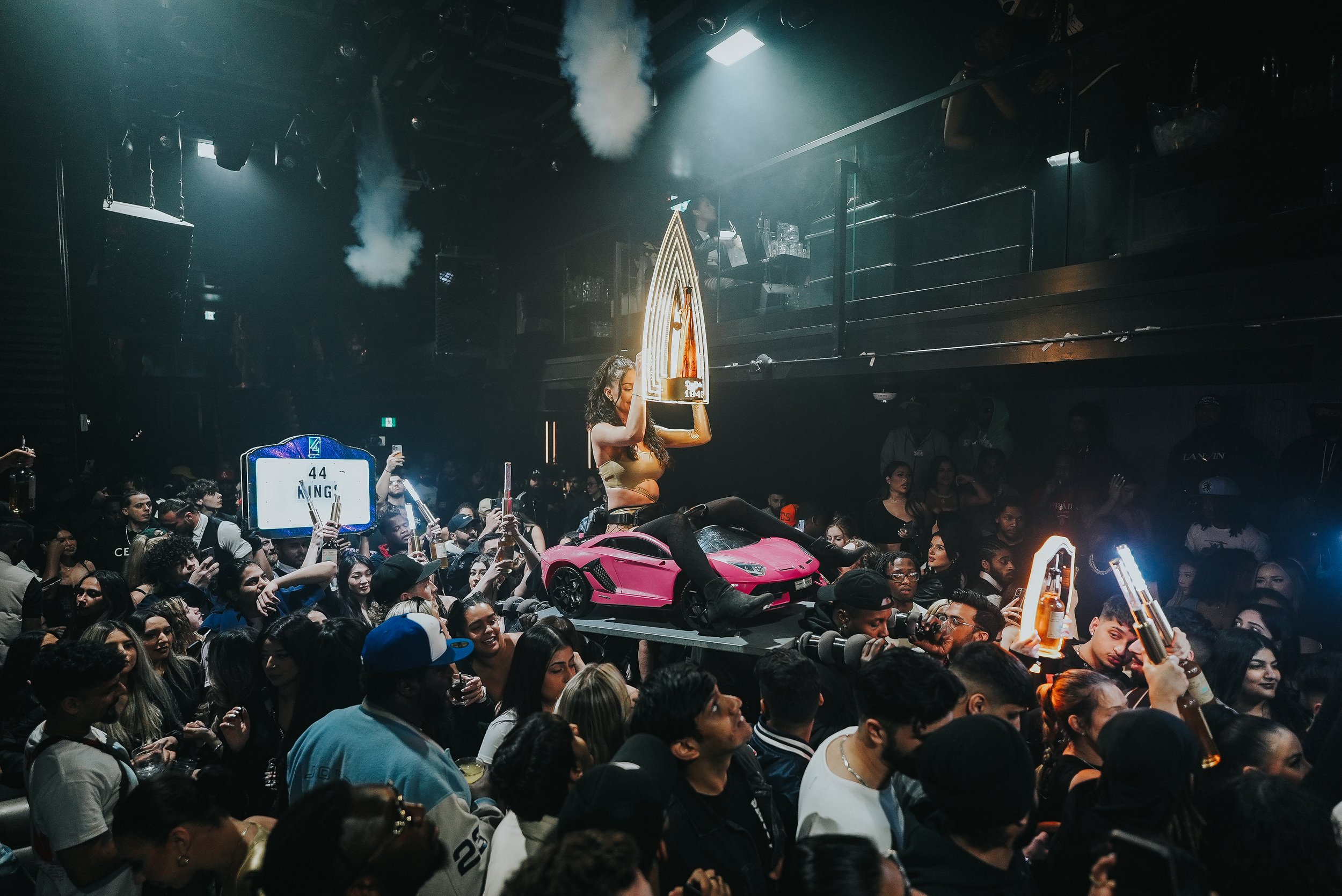 A crowded nightclub scene with a female performer holding a glass award, surrounded by fans, with a pink toy Lamborghini in the foreground and DJ equipment on an elevated stage in the background.