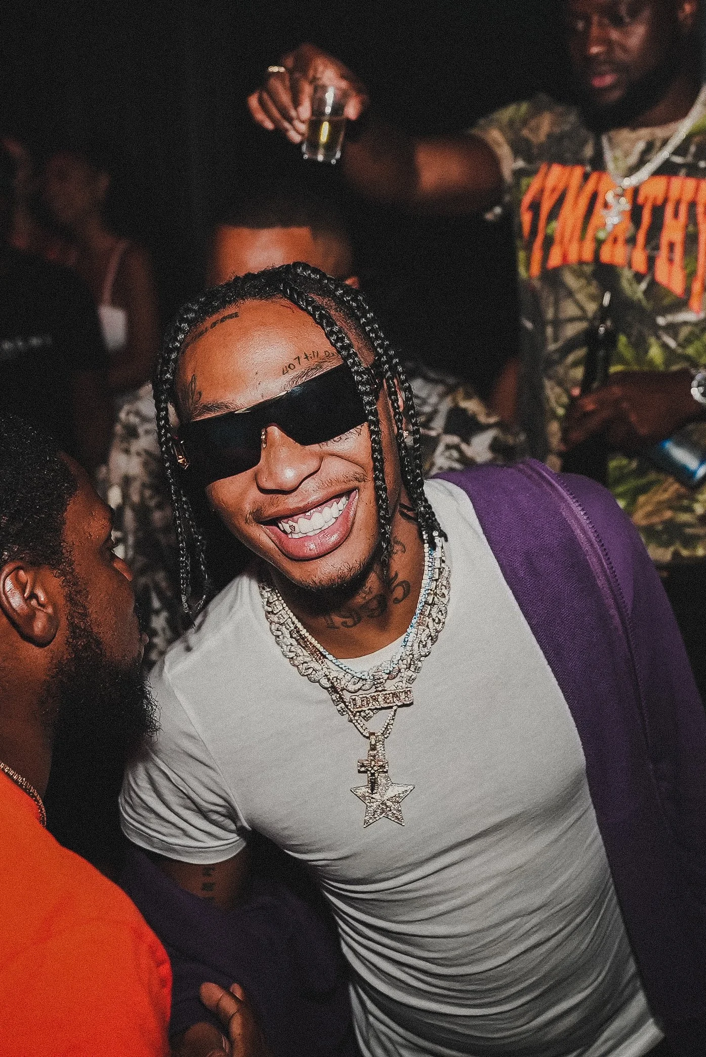 Close-up of a man with braided hair, wearing sunglasses, a white t-shirt, and a large, silver, star-shaped chain necklace, smiling at a social gathering.