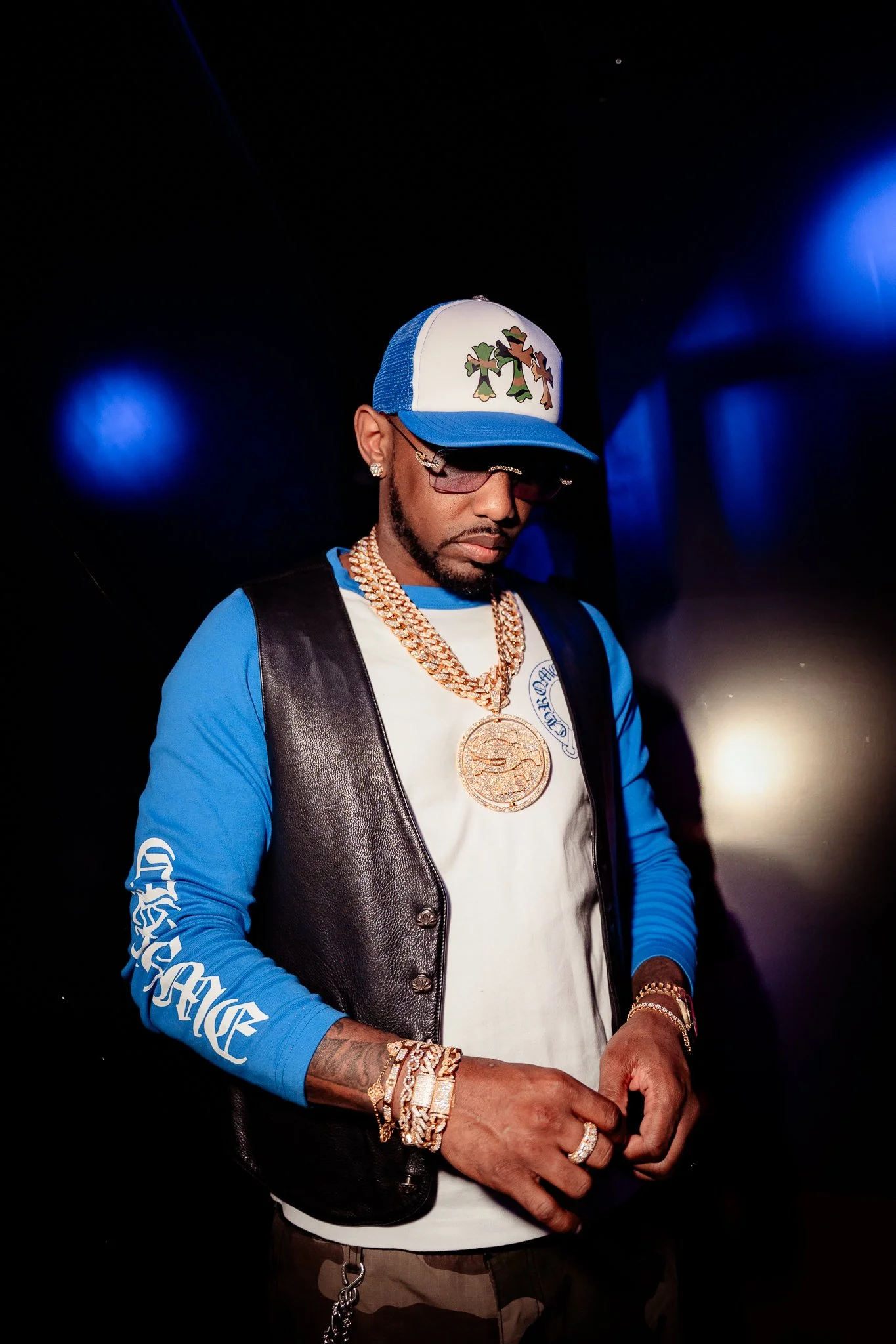 Man wearing a blue and white trucker hat with cartoon characters, glasses, gold chains, earrings, and multiple bracelets, standing in a dark setting with blue lights.